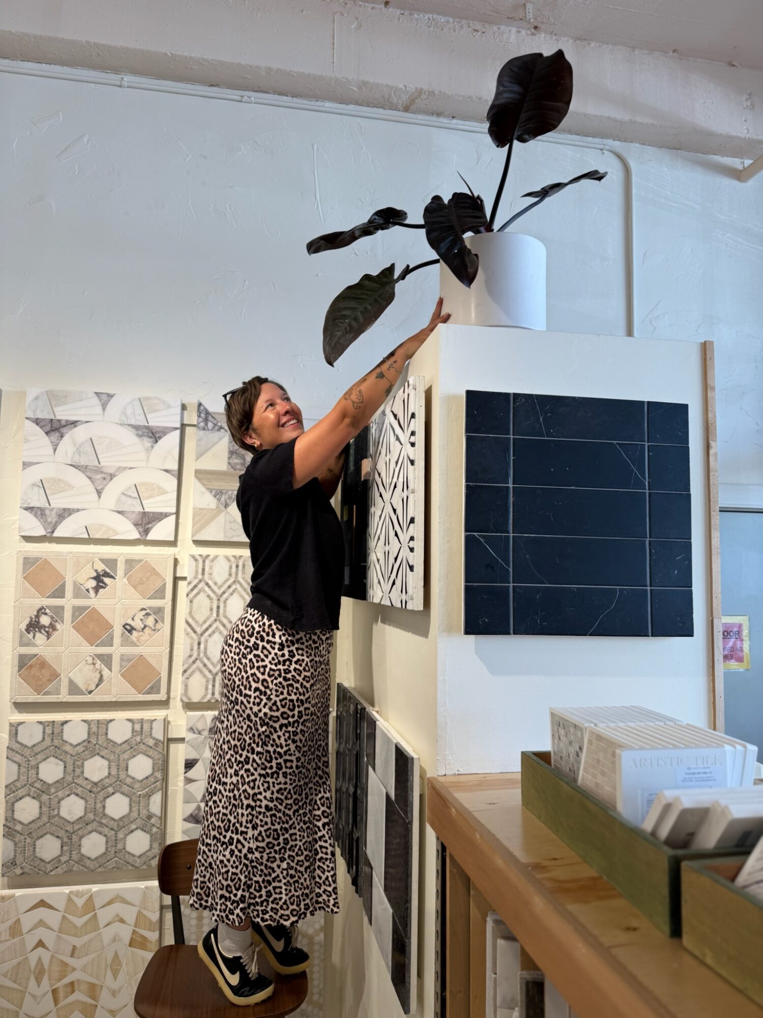 Woman reaching up to a potted plant on a high shelf, with decorative tiles and a display of tiles in the background.