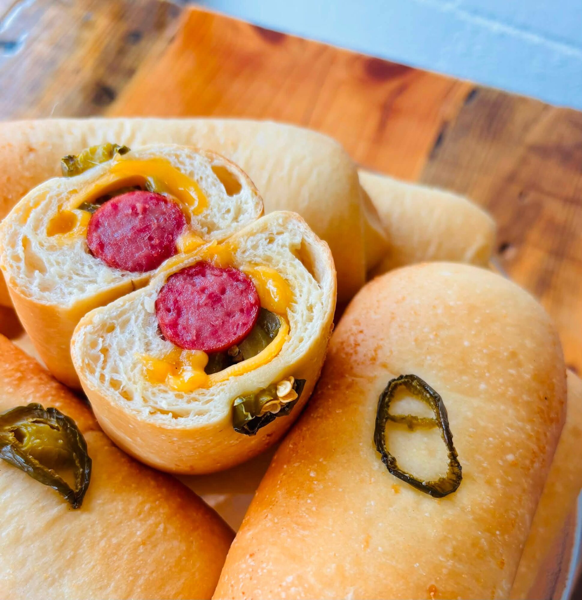 Close-up of bread rolls with fillings, one cut open showing sausage, cheese, and vegetables inside, on a wooden surface.