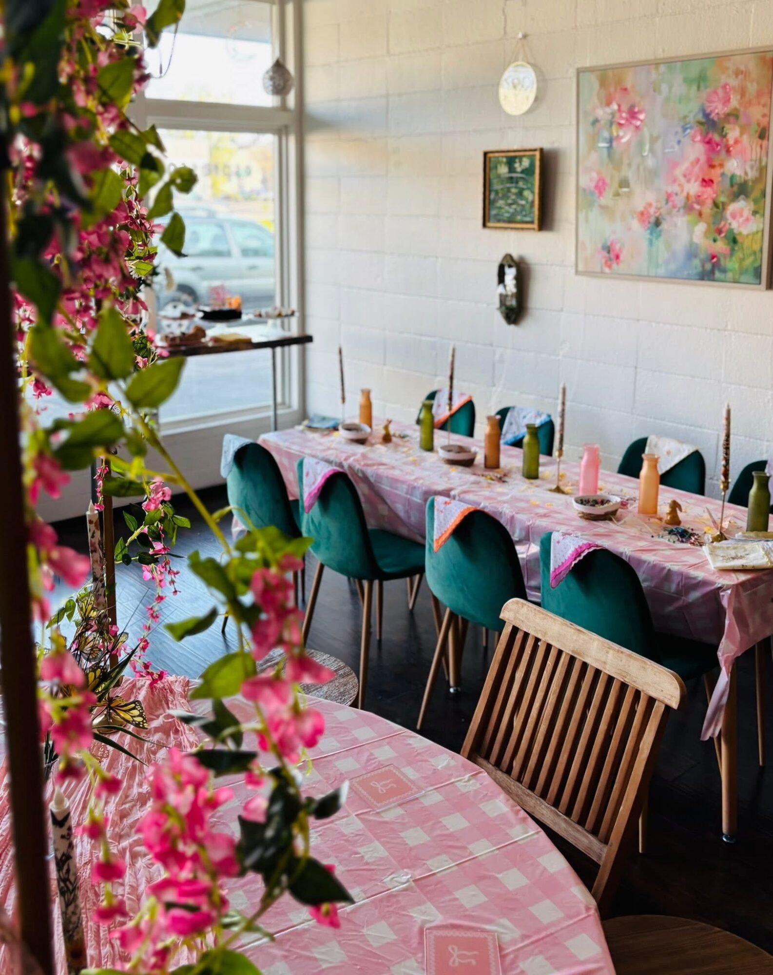 Long table set for a celebration with pink tablecloth, candles, and chairs, decorated with flowers and artwork on the wall.
