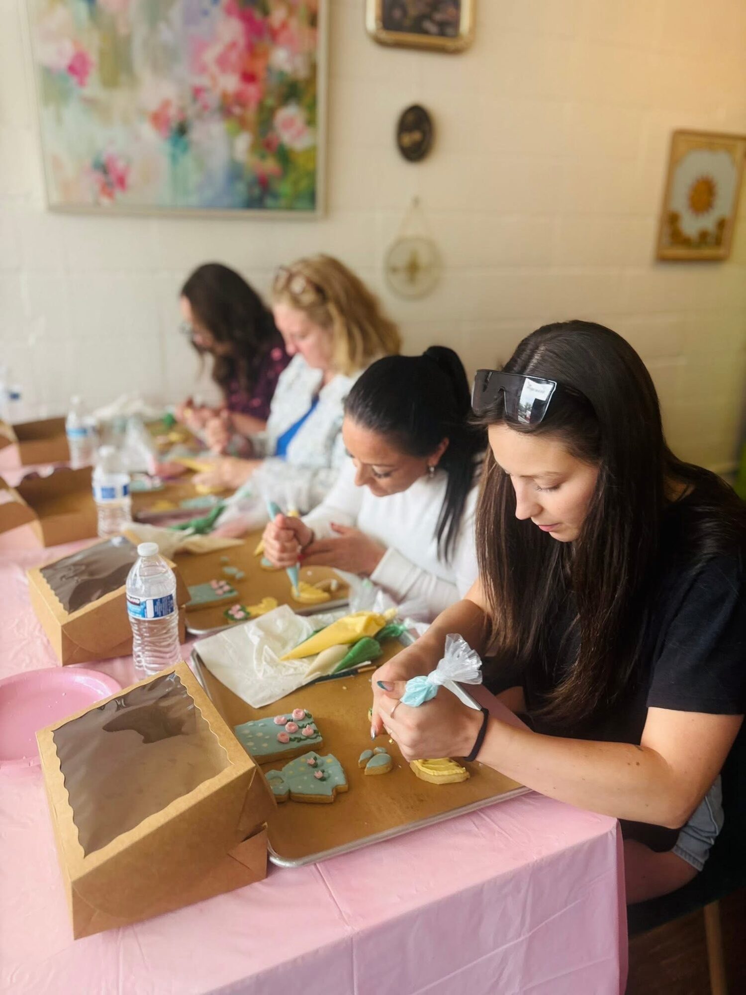 Four women sit at a table working on craft projects, with art on the wall behind them.