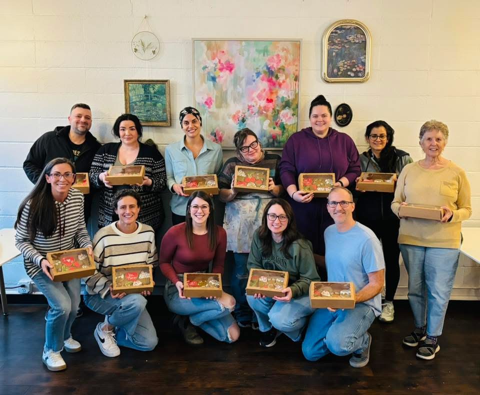 Group of twelve people holding boxes, standing and kneeling indoors with artwork on the wall behind them.