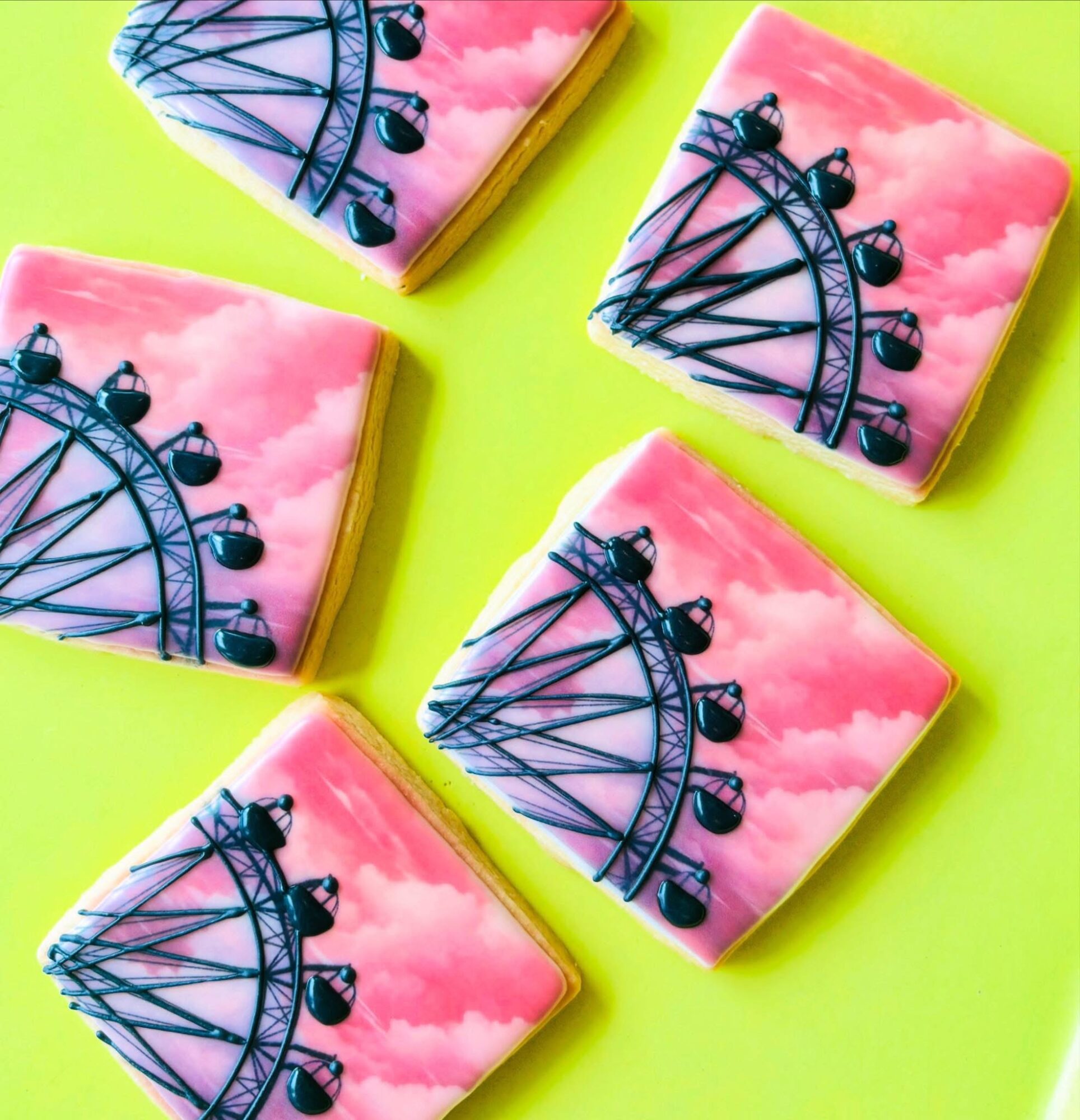 Four square cookies with pink cloud backgrounds and a black Ferris wheel design, on a bright green surface.