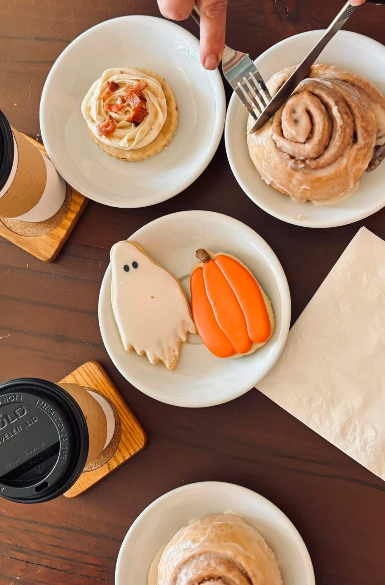 Two decorated cookies, one ghost and one pumpkin, on a white plate, with drinks and food on a dark table.
