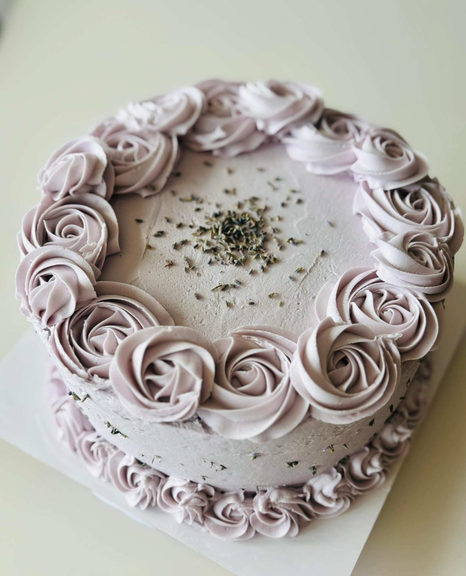 Round cake decorated with light pink roses around the top edge and sprinkled with small green herbs or seeds in the center.