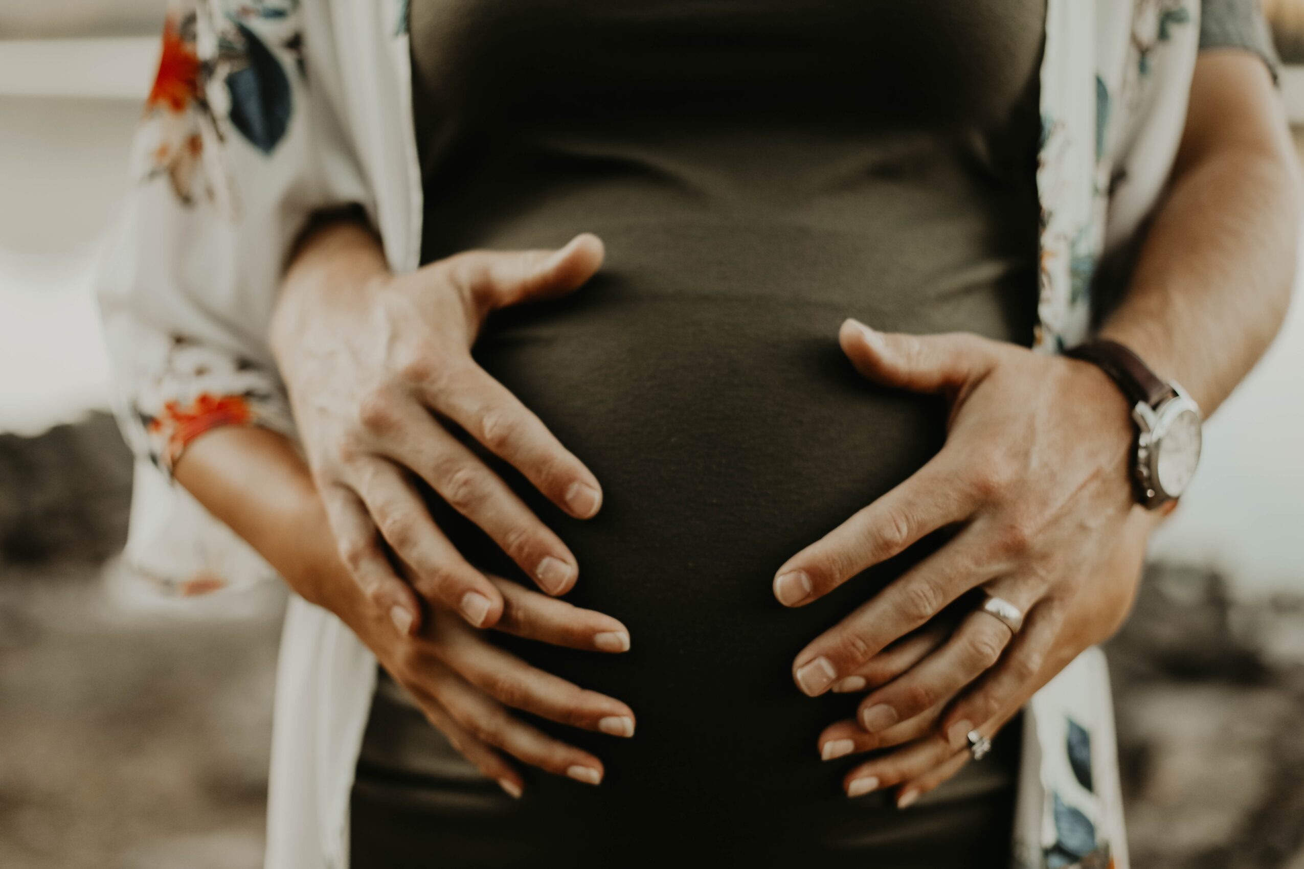 Close-up of a pregnant person's belly with hands gently resting on it, wearing a watch and ring, outdoors.