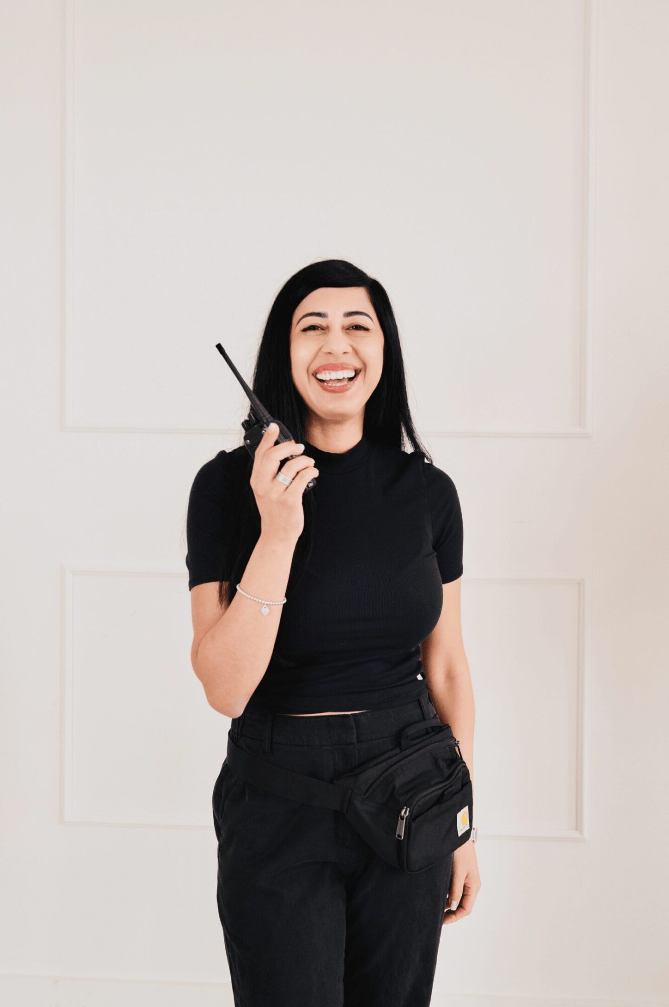 Young woman smiling and holding a walkie-talkie, wearing a black shirt and pants, standing against a plain background.