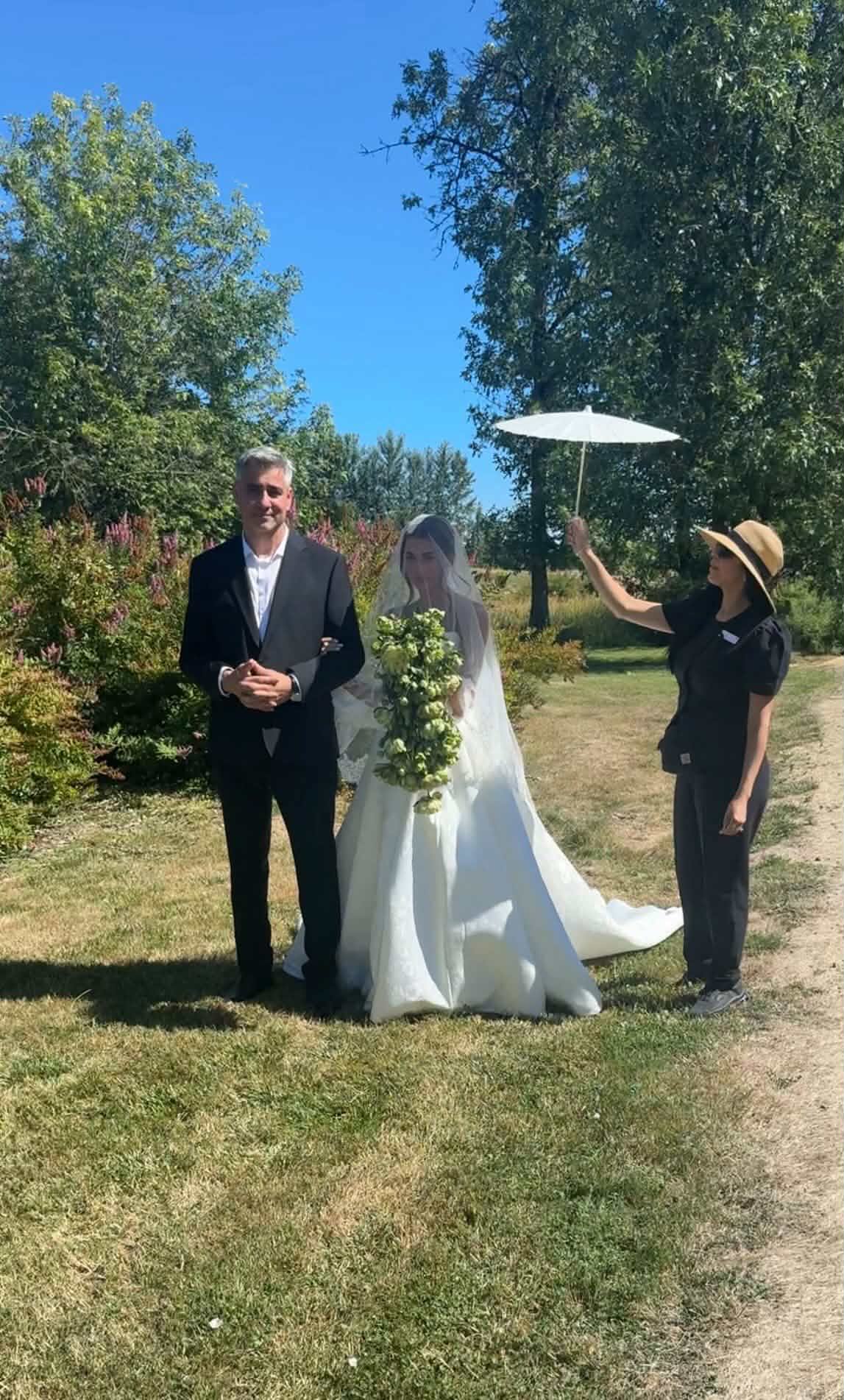 Three people outdoors: a man in a suit, a woman in a wedding dress holding a bouquet, and a woman with a hat holding an umbrella, on a sunny day with trees.