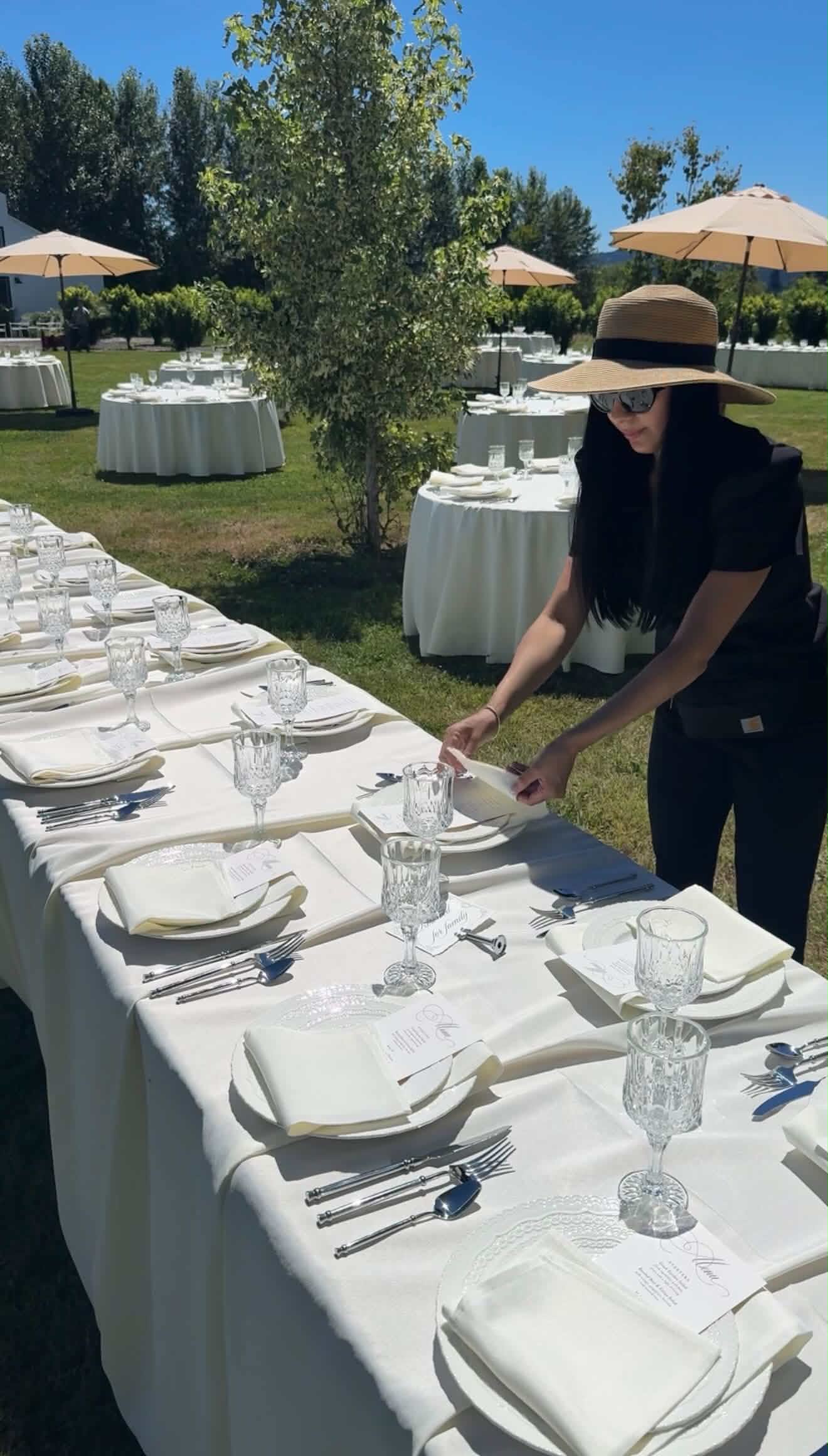 Person setting a long outdoor table with white tablecloths, glasses, and plates, under umbrellas on a sunny day.