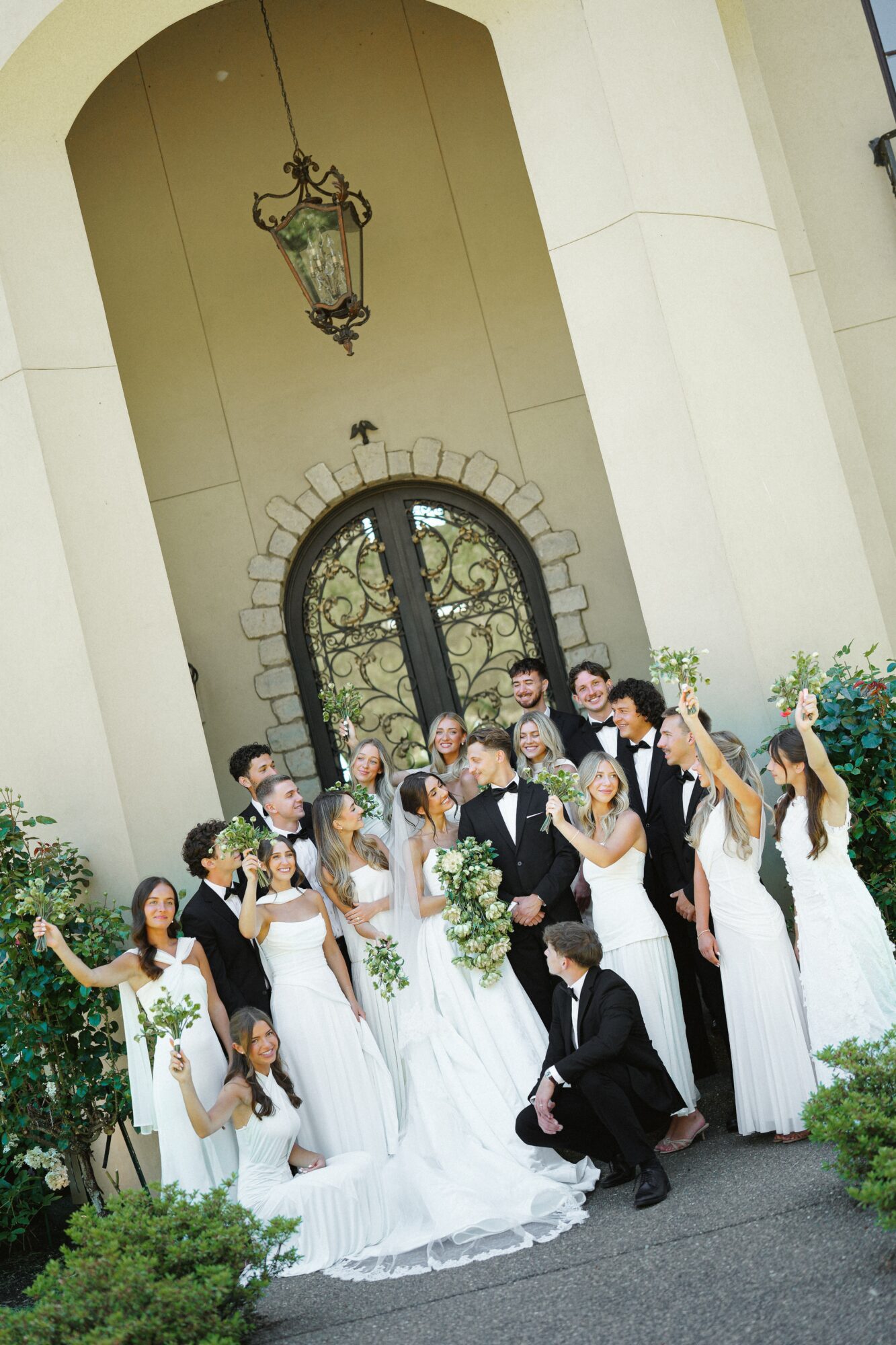 Group of people in wedding attire outside building with arched door and hanging lantern, smiling and posing for photo.