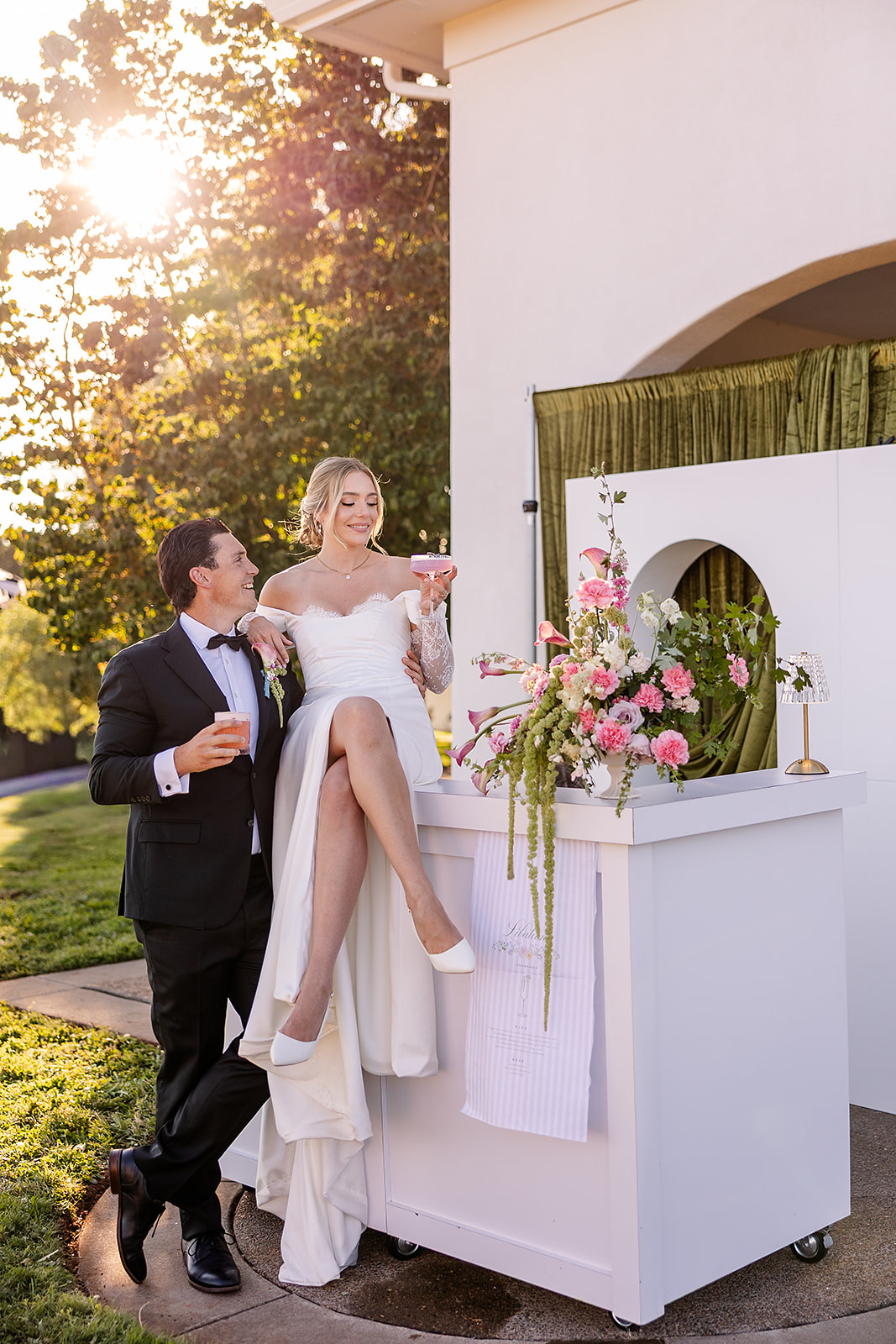 Bride in a white wedding dress and groom in a tuxedo stand outdoors near a white structure with a green curtain, sunlight shining through trees.