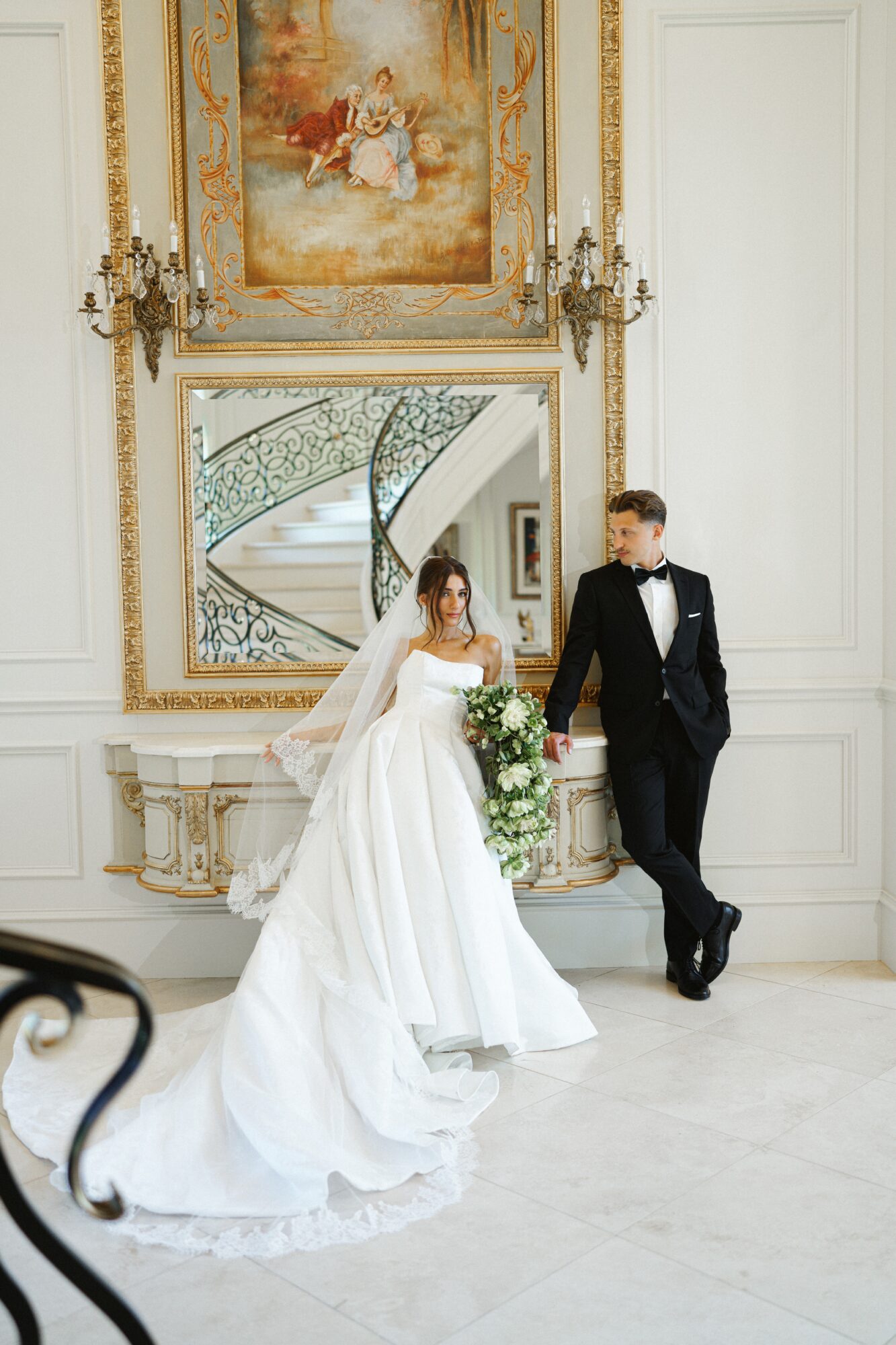 Bride and groom in elegant wedding attire in a luxurious room with ornate decor and a large mirror.