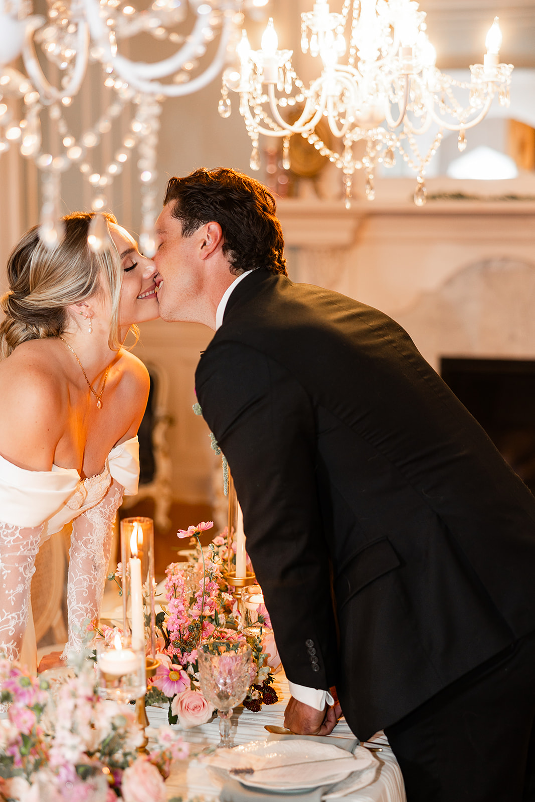 A couple kisses at a wedding reception with chandeliers and floral arrangements, woman in orange dress, man in black suit.
