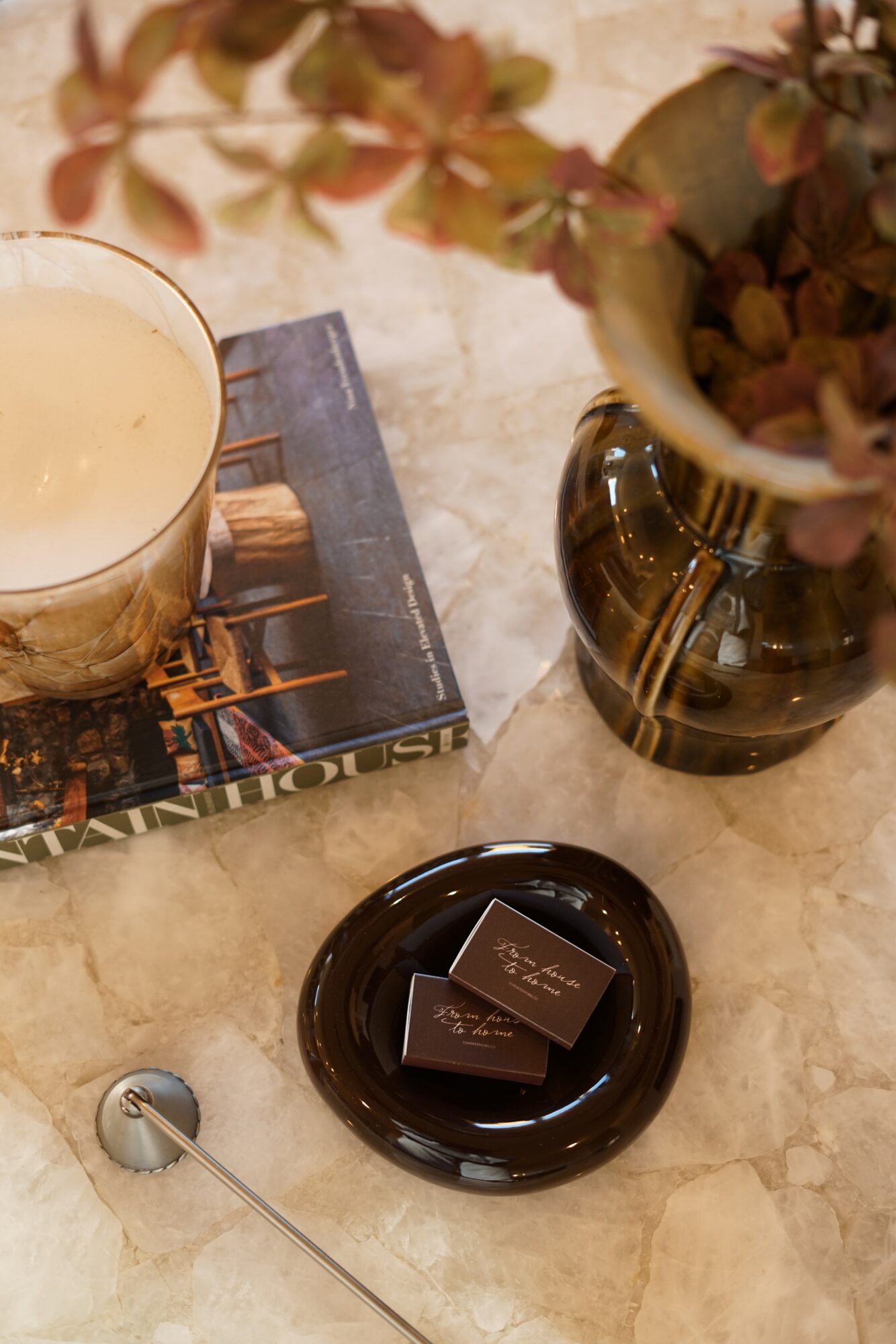 Vase with dried flowers, candle on a book, and chocolates on a plate on a marble surface.