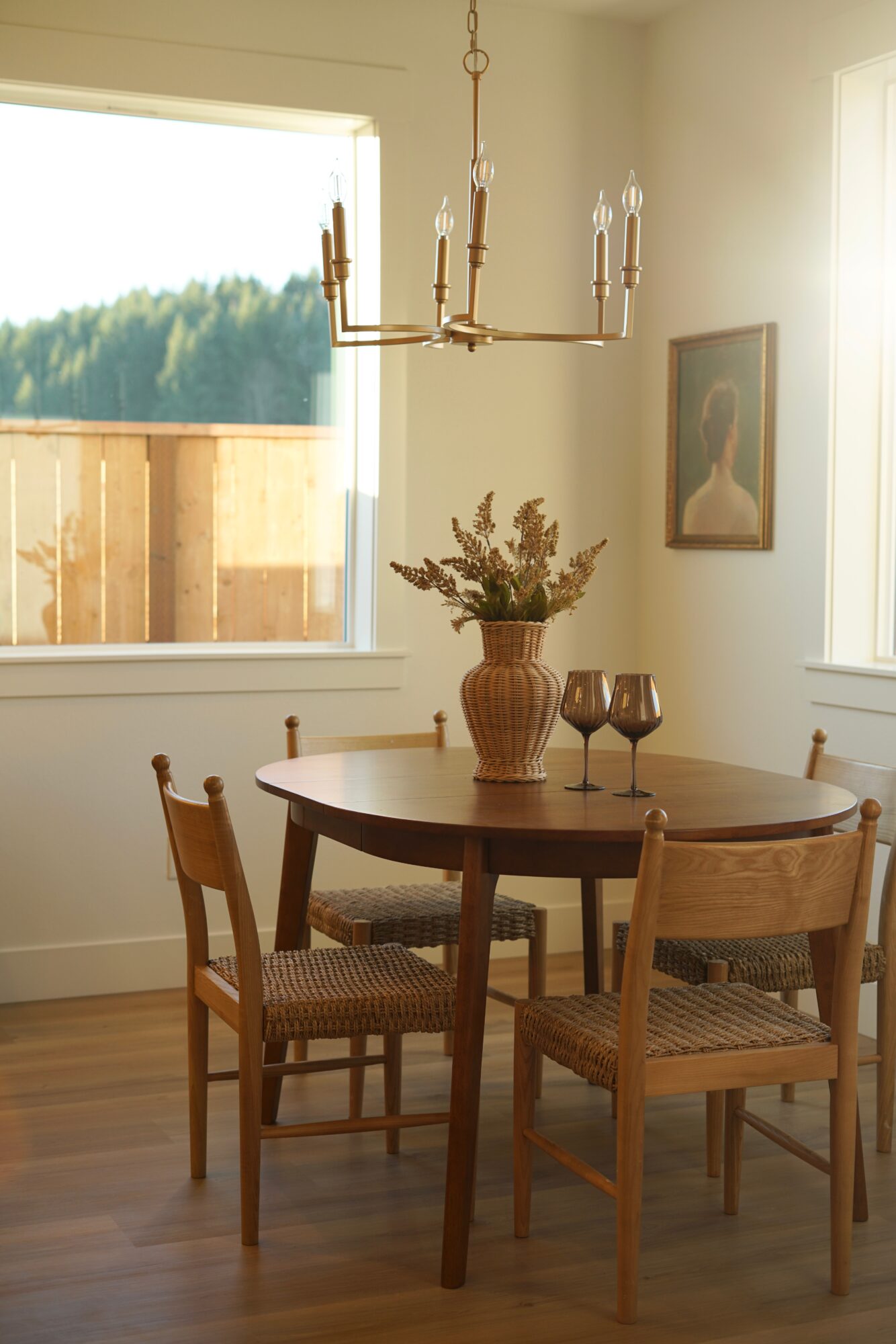 Dining room with round wooden table, four chairs, a vase with flowers, two wine glasses, chandelier, and window with view.