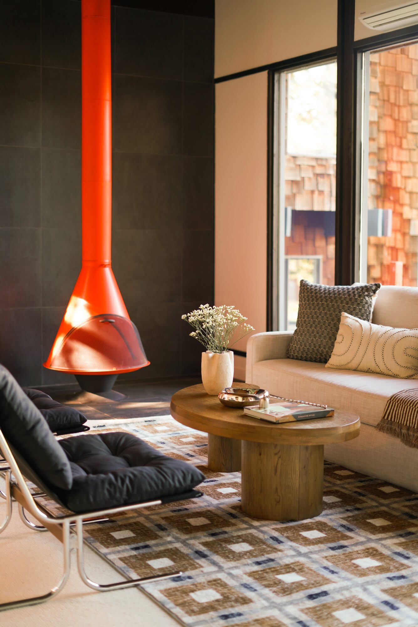 Living room with a red hanging lamp, beige sofa, round wooden coffee table, and a patterned rug, near large windows.
