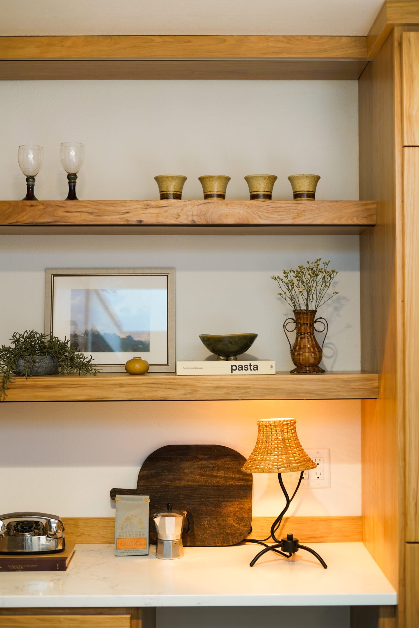 Wooden shelves with glassware, bowls, framed picture, plant, and a lamp on a white surface.