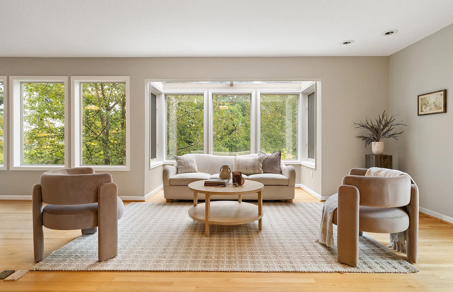 Living room with large windows, two armchairs, a sofa, a coffee table, and a rug, with a plant and framed picture.