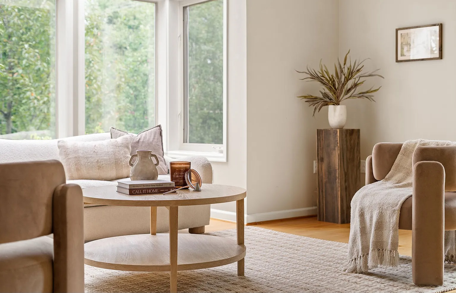 Living room with large windows, sofa, armchair, round coffee table, and a tall plant on a wooden stand.