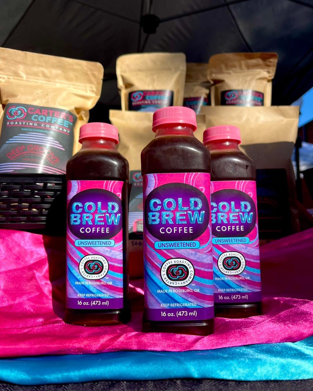 Three bottles of cold brew coffee with pink caps and colorful labels in front of brown paper bags, on a pink and blue surface.