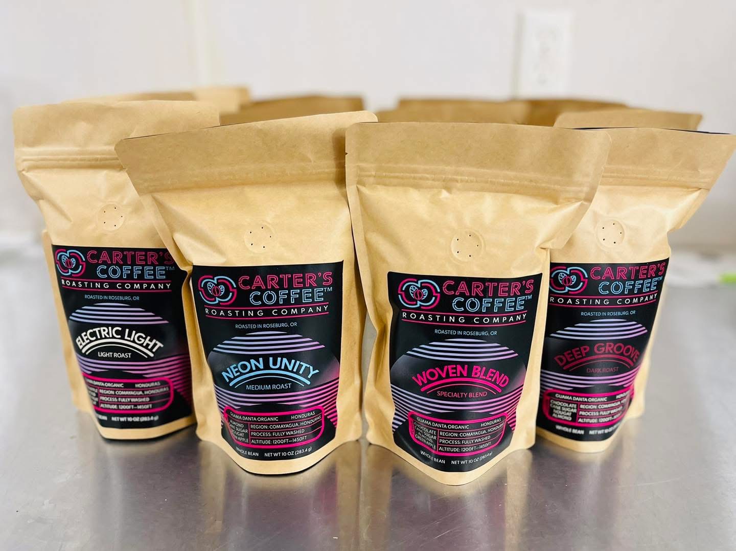 Six brown coffee bags with colorful labels on a gray surface, background is white wall with electrical outlet.