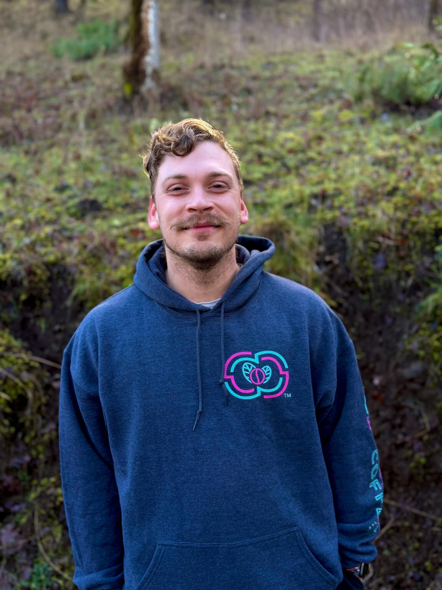 Young man with short curly hair and a beard, smiling, wearing a blue hoodie with a colorful logo, outdoors in a natural setting.