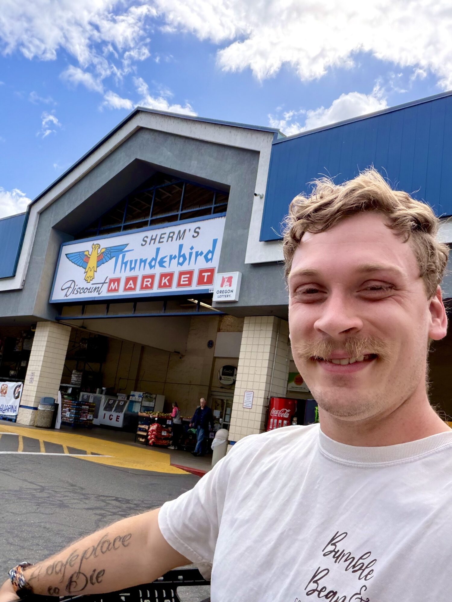 Young man with curly hair smiling in front of a building with a sign reading 'Sherm's Thunderbird Market' under a partly cloudy sky.