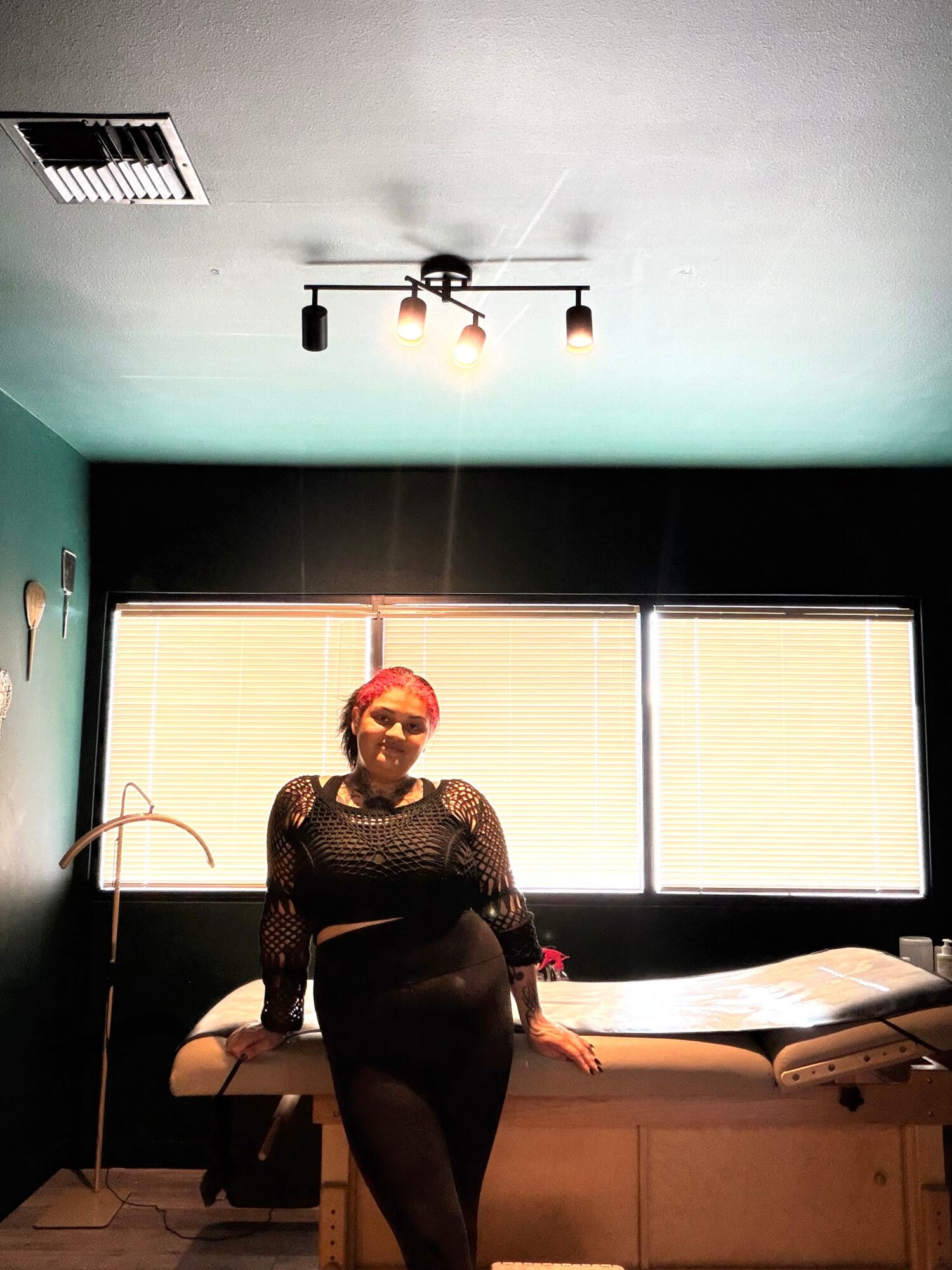 Woman with red hair and glasses standing in a room with large windows, a medical examination table, and a ceiling light.