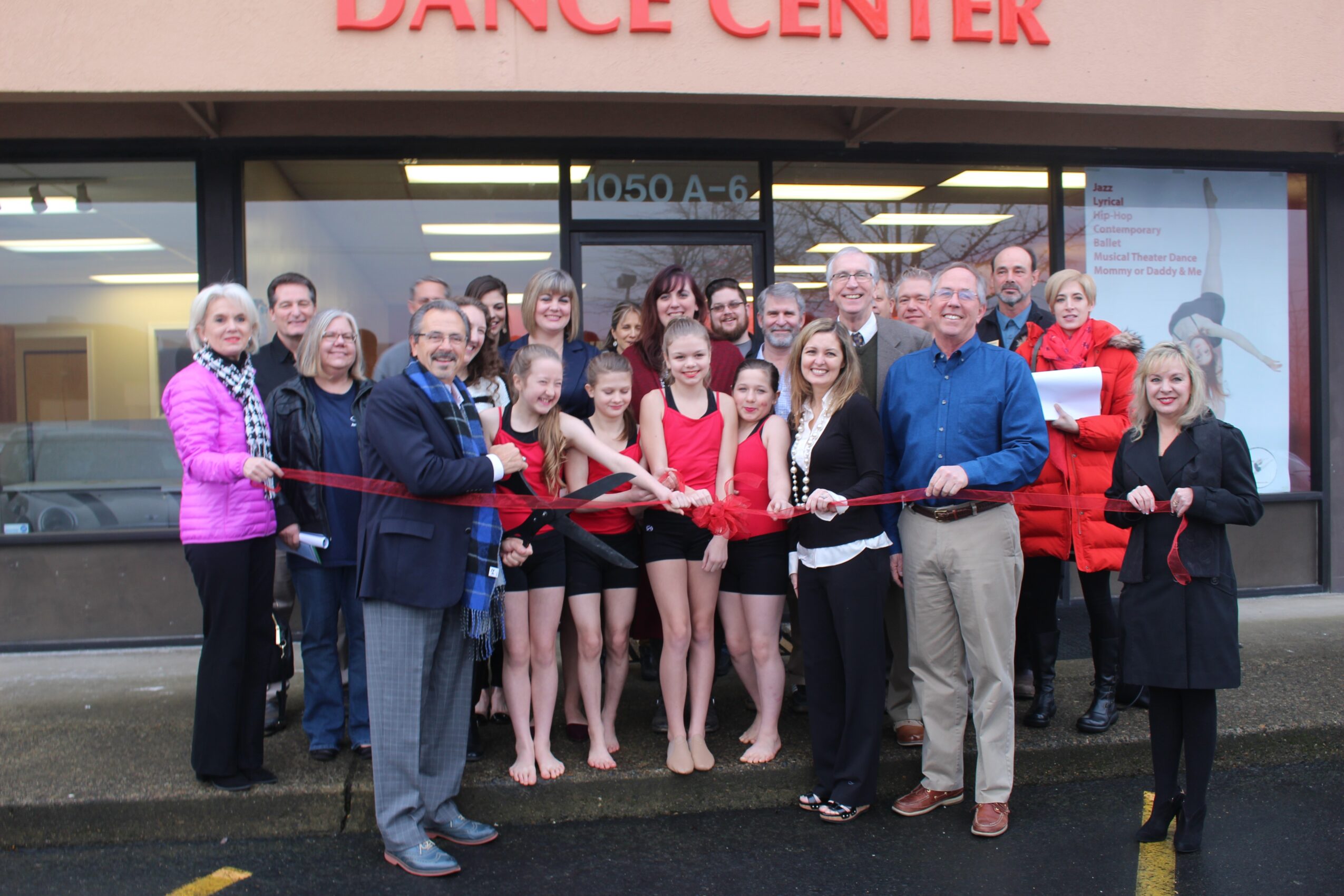 Group of people standing outside a dance center, some holding a ribbon for a ribbon-cutting event, in front of a building with large windows.