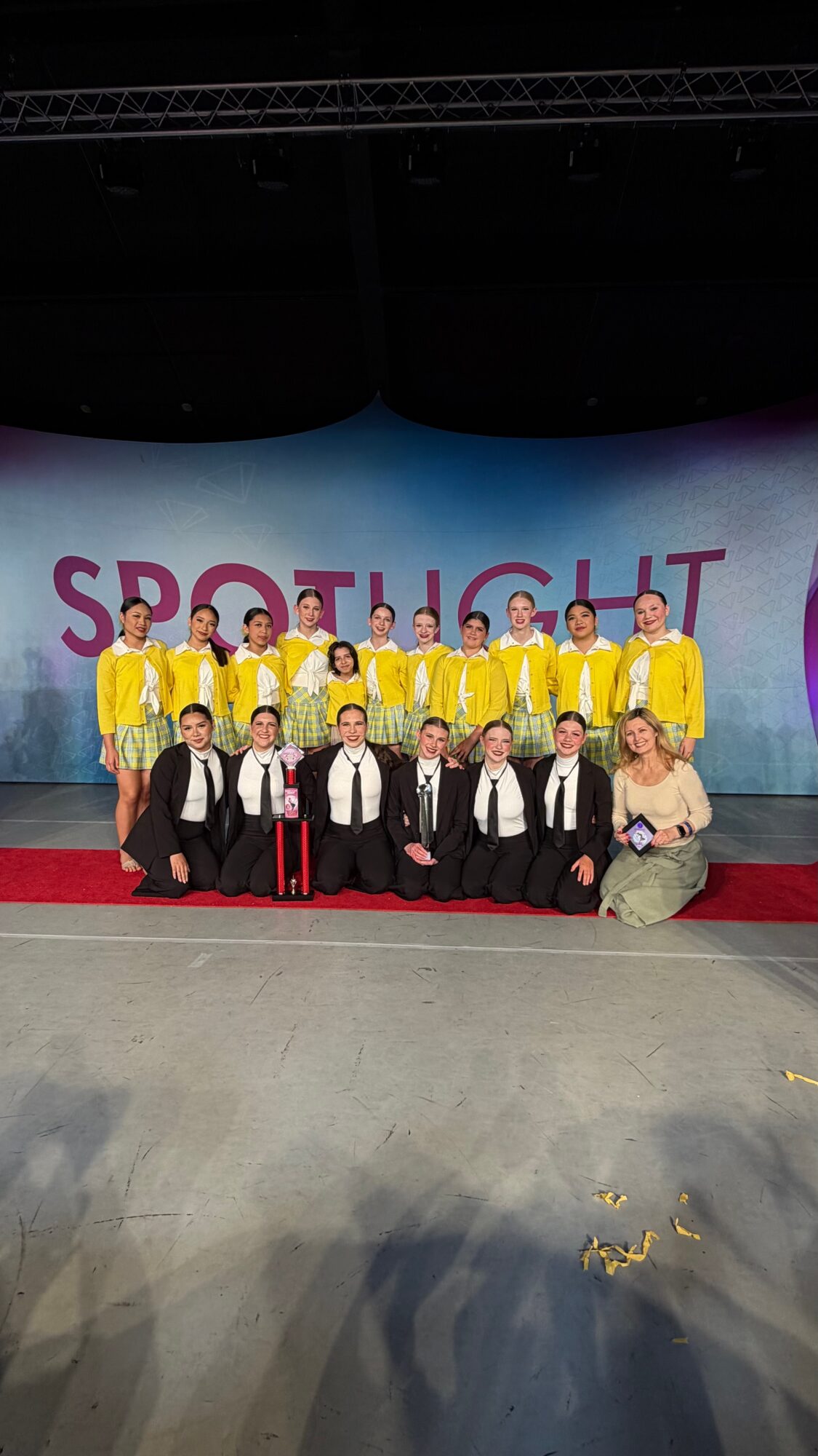 Group of children in yellow costumes and adults in formal attire on stage with a large screen behind them displaying 'SPOTLIGHT'.