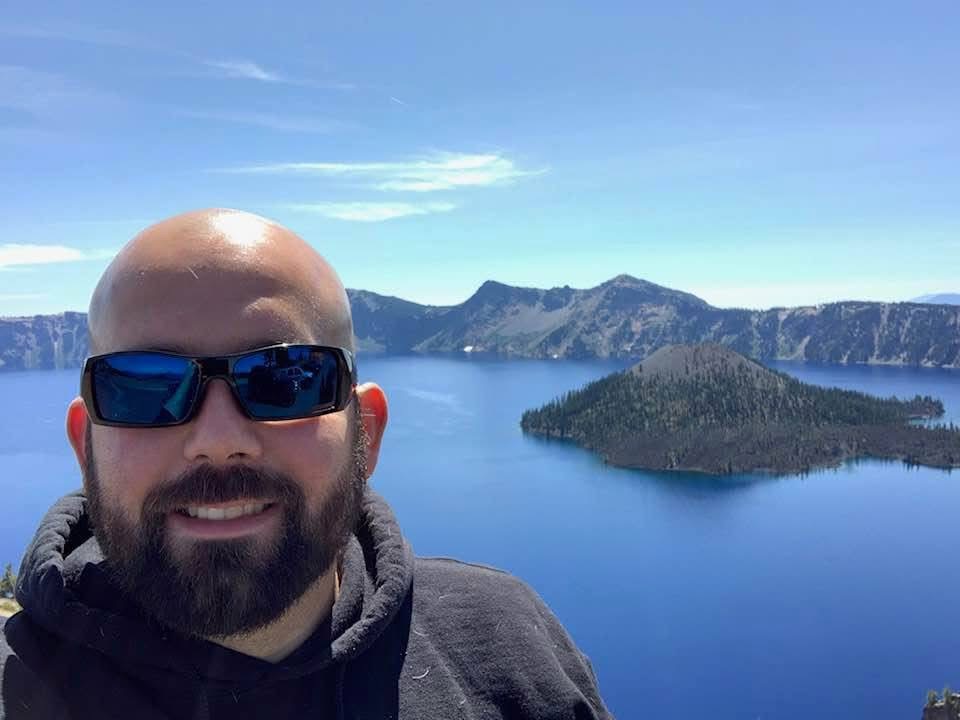 Bald man with sunglasses smiling outdoors with a lake and mountains in the background.