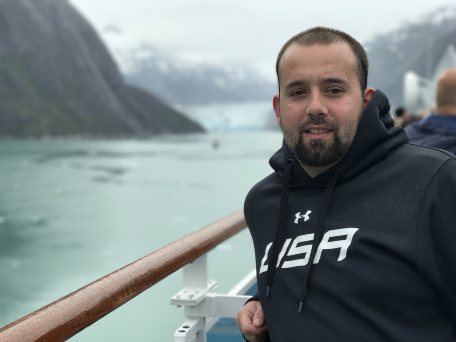 Man with beard in black hoodie on boat with mountains and water in background