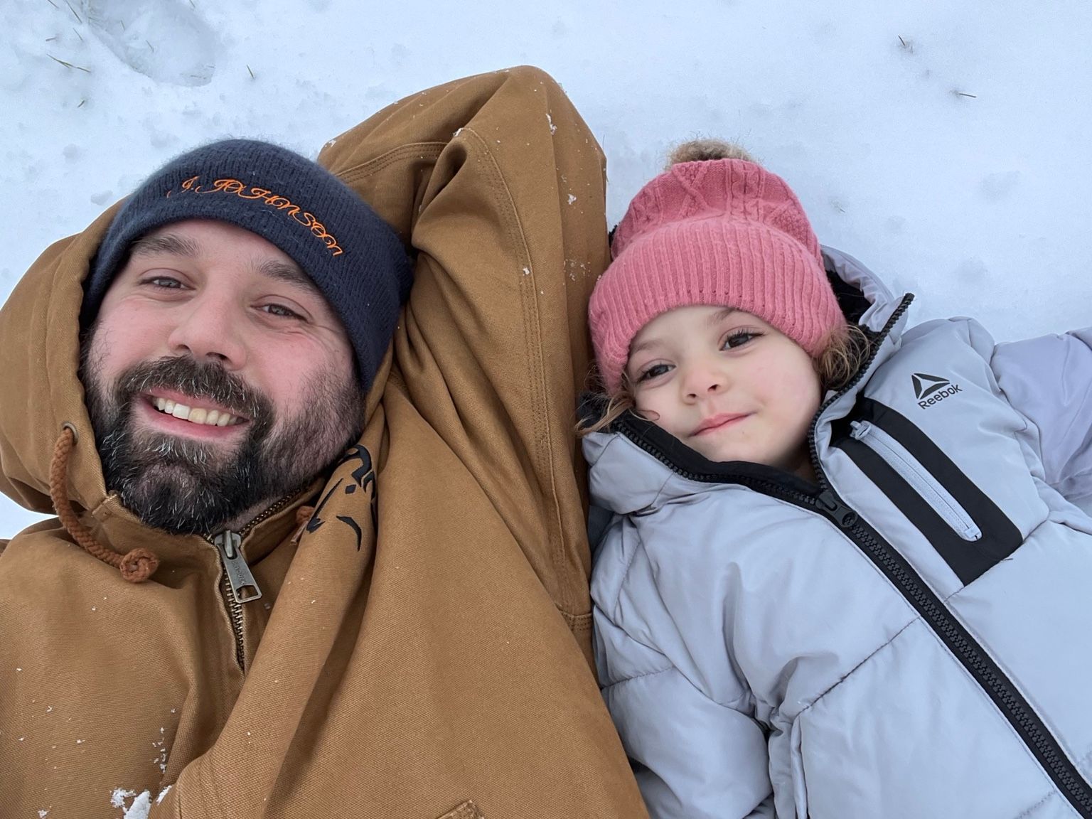 A woman and a man lying on snow, smiling, with the woman wearing a pink hat and gray jacket, and the man in a brown jacket and black beanie.