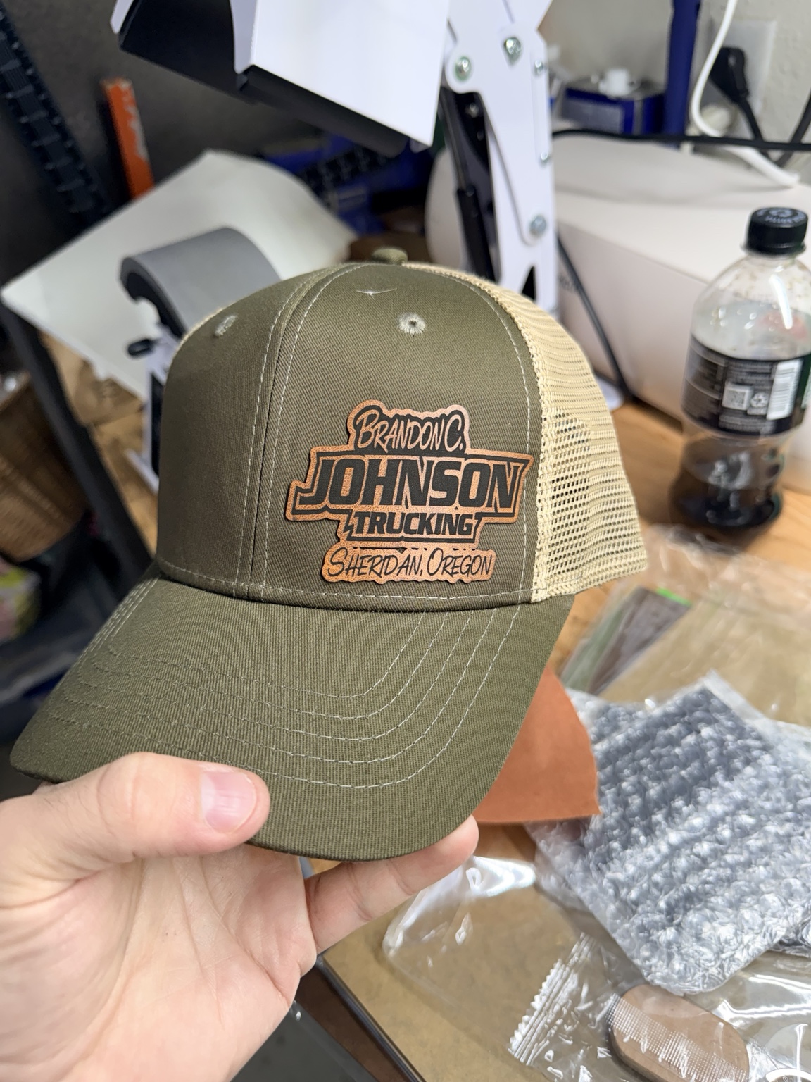 Olive green trucker cap with embroidered patch reading 'Brandon Johnson Trucking' held by a hand, cluttered workspace background.