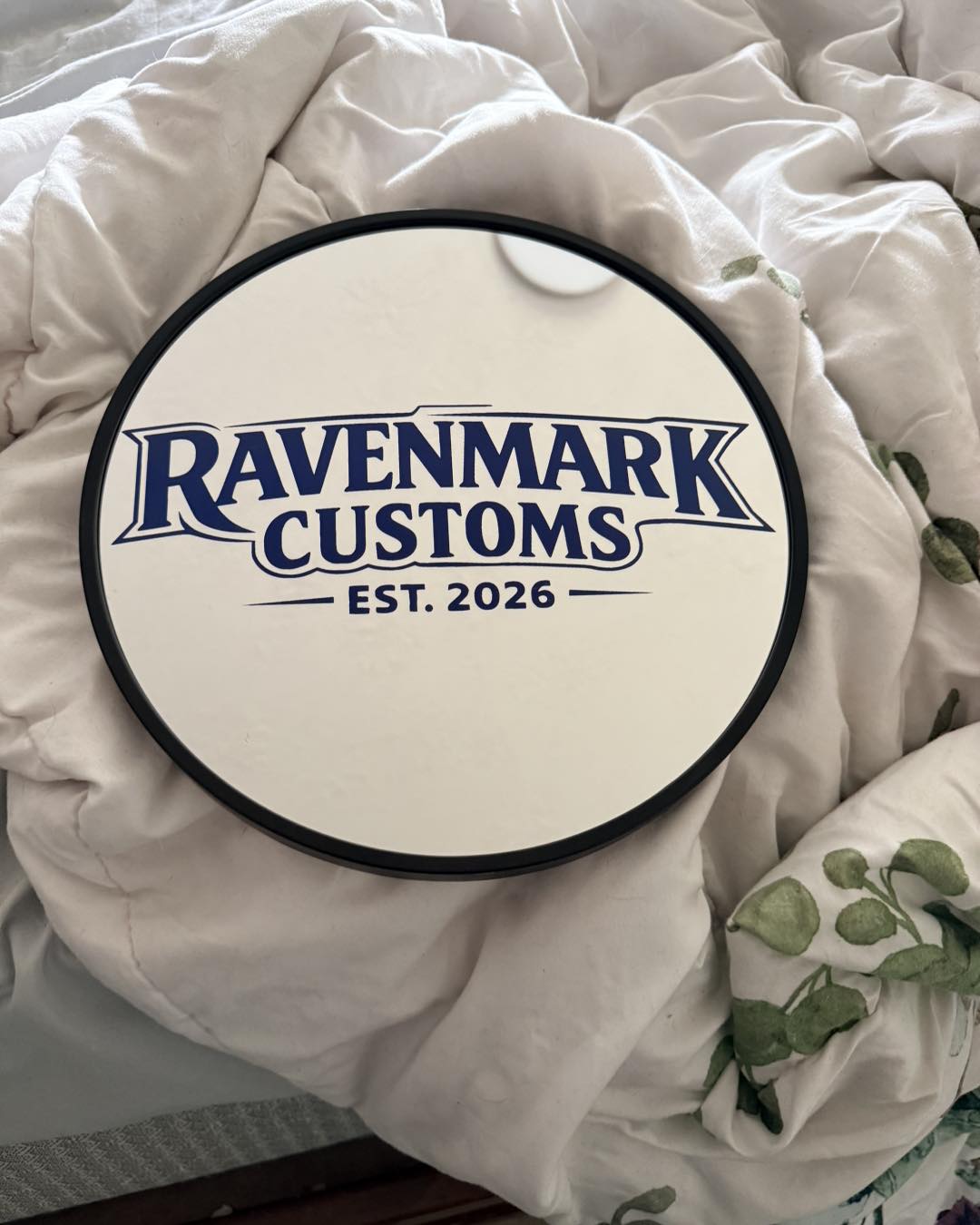 Round sign with blue text reading 'Ravenmark Customs EST. 2026' on a white background, placed on a bed with white bedding and green leaves.