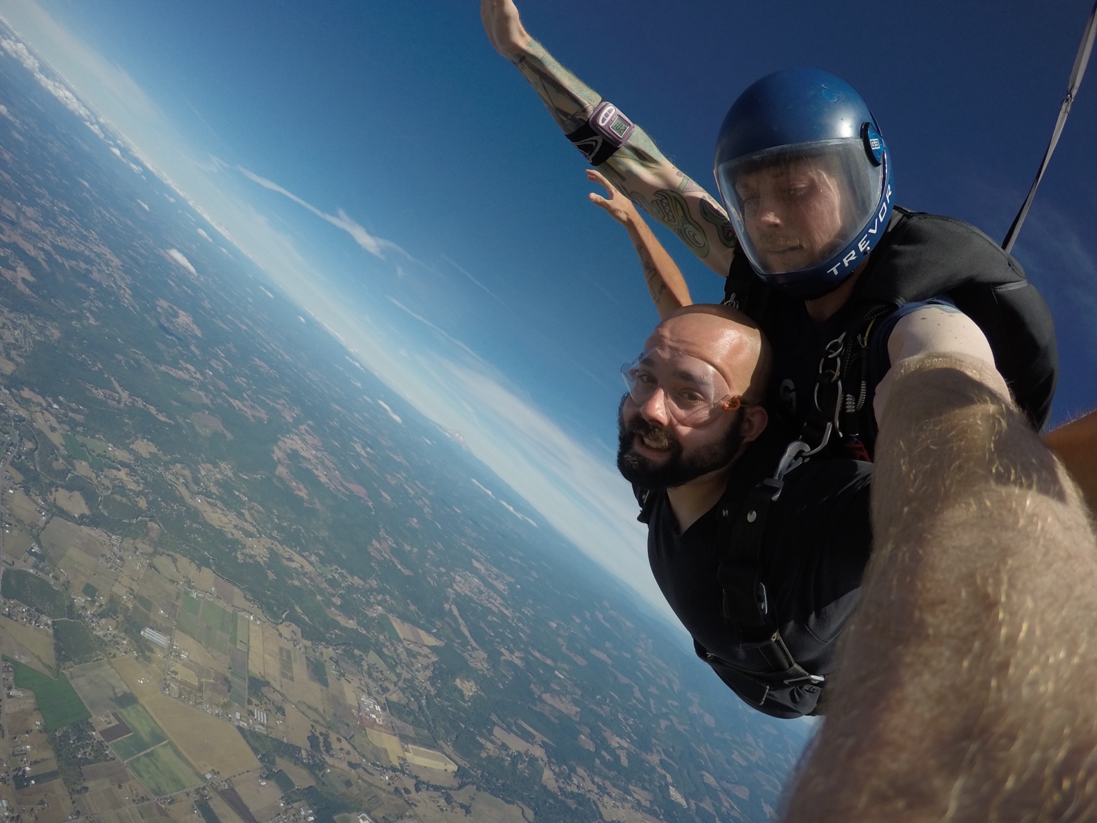 Two skydivers free-falling above land with fields and sky visible, one taking a selfie with a camera, wearing helmets and jumpsuits.
