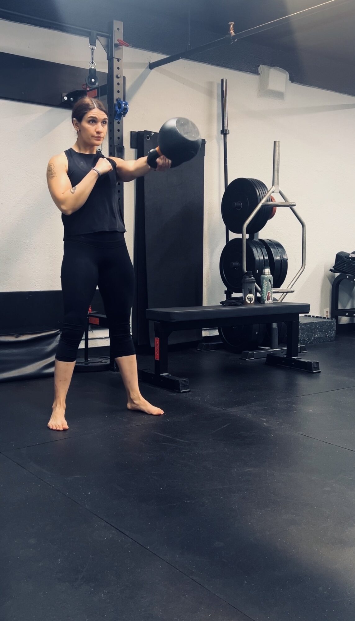 Woman holding a medicine ball in a gym, standing barefoot on black flooring, with gym equipment in background.