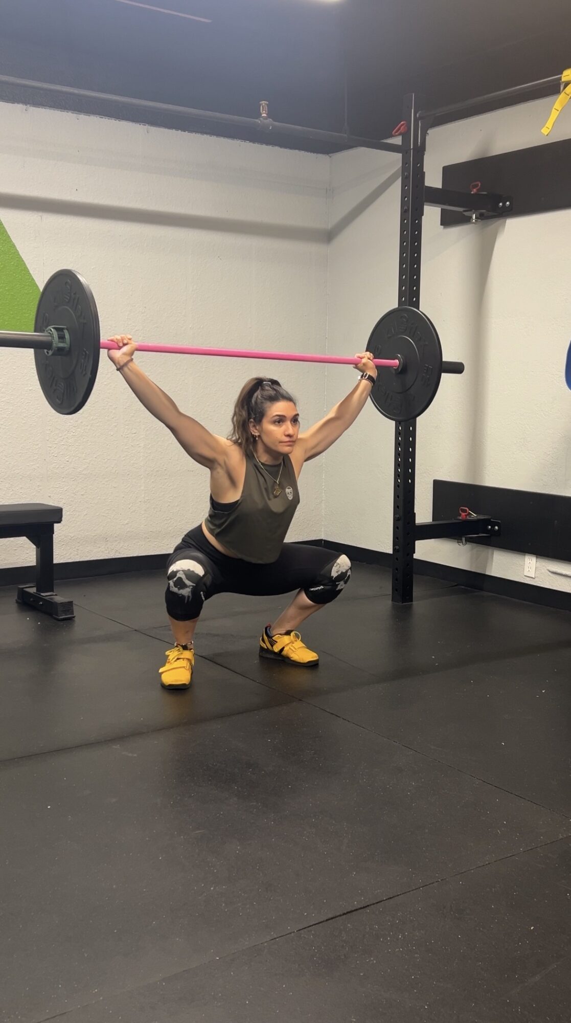 Woman in squat position holding a barbell overhead in a gym, with black flooring and equipment in background.