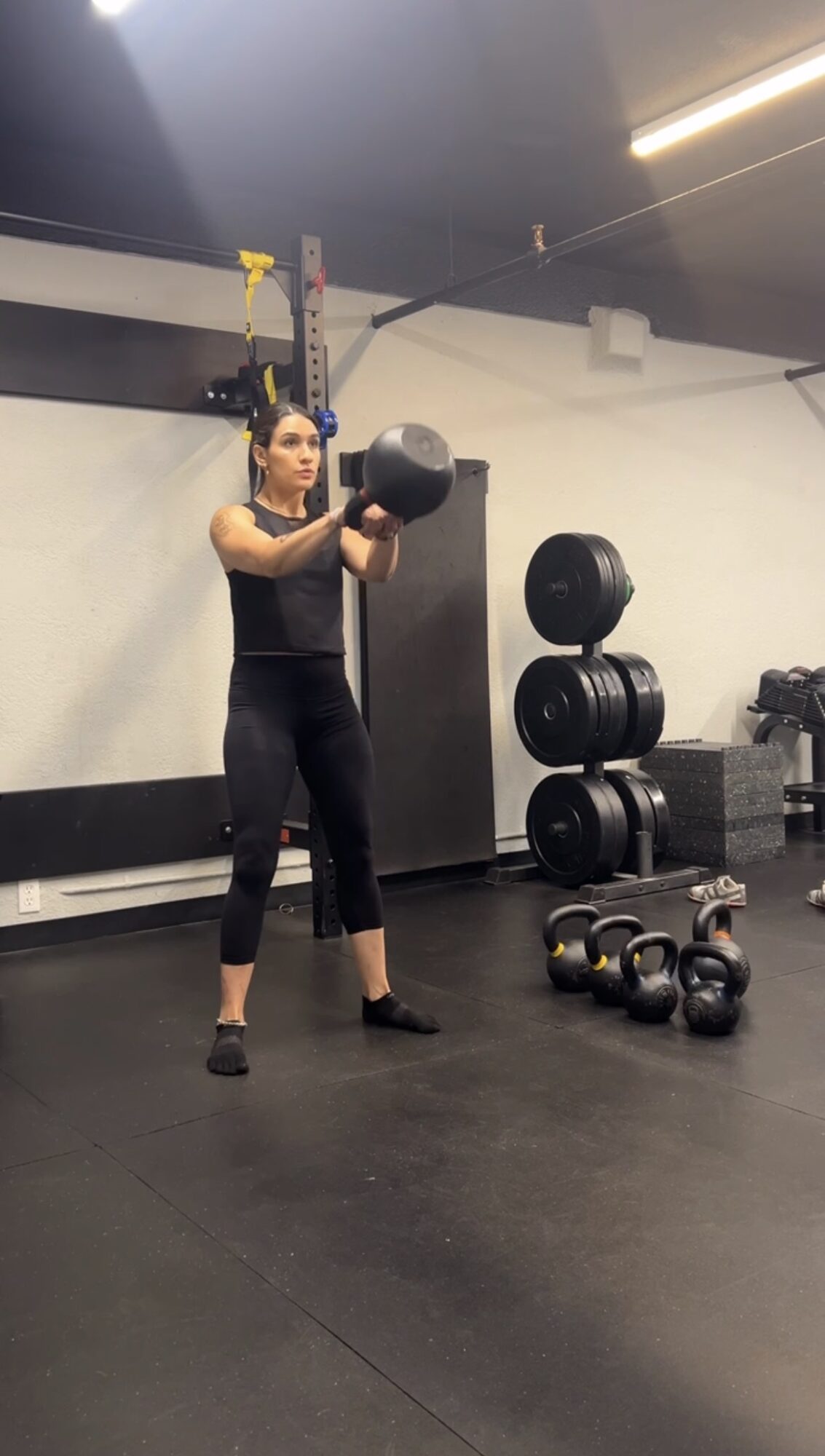Woman in black workout clothes holding a medicine ball in a gym, with kettlebells and stacked weights nearby.
