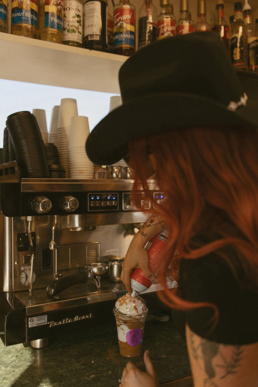 Person with long hair and a hat preparing coffee behind a bar, with cups and bottles in the background.