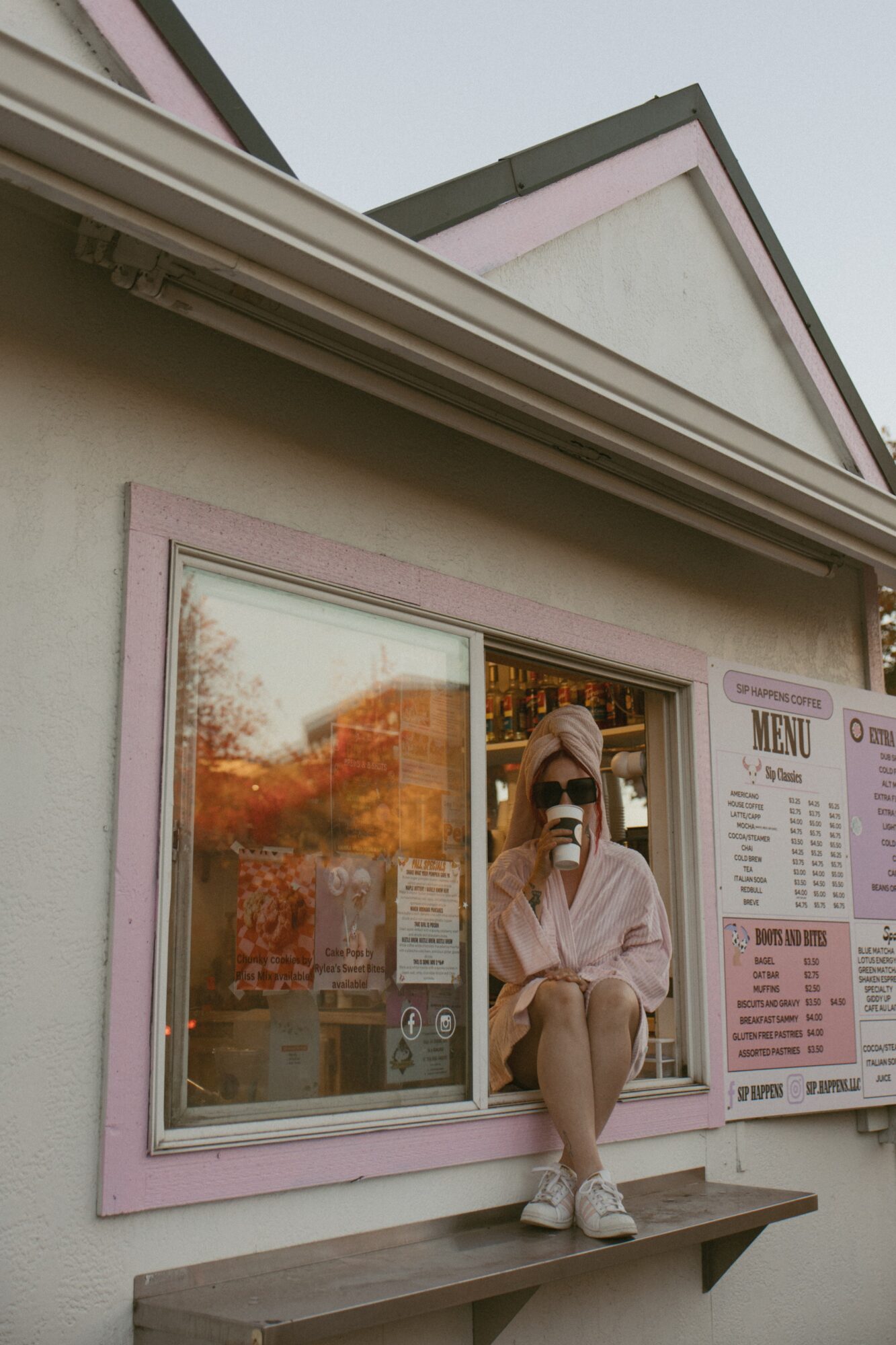 Person sitting on window ledge taking a photo with a phone, reflected in window, building exterior with pink trim, menu board nearby.