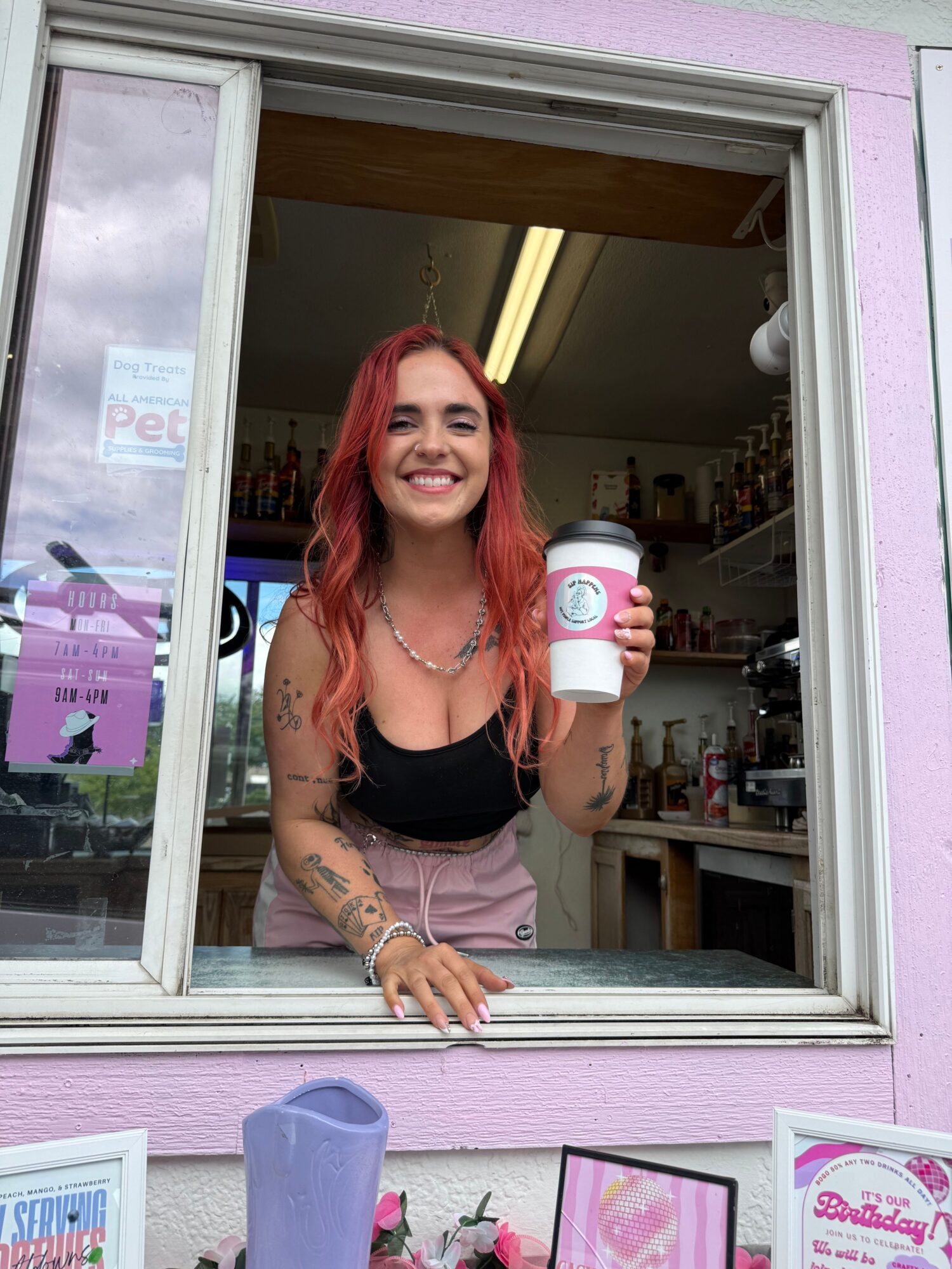 Woman with pink hair holding a bottle, smiling, standing at a store counter with pink walls and products.