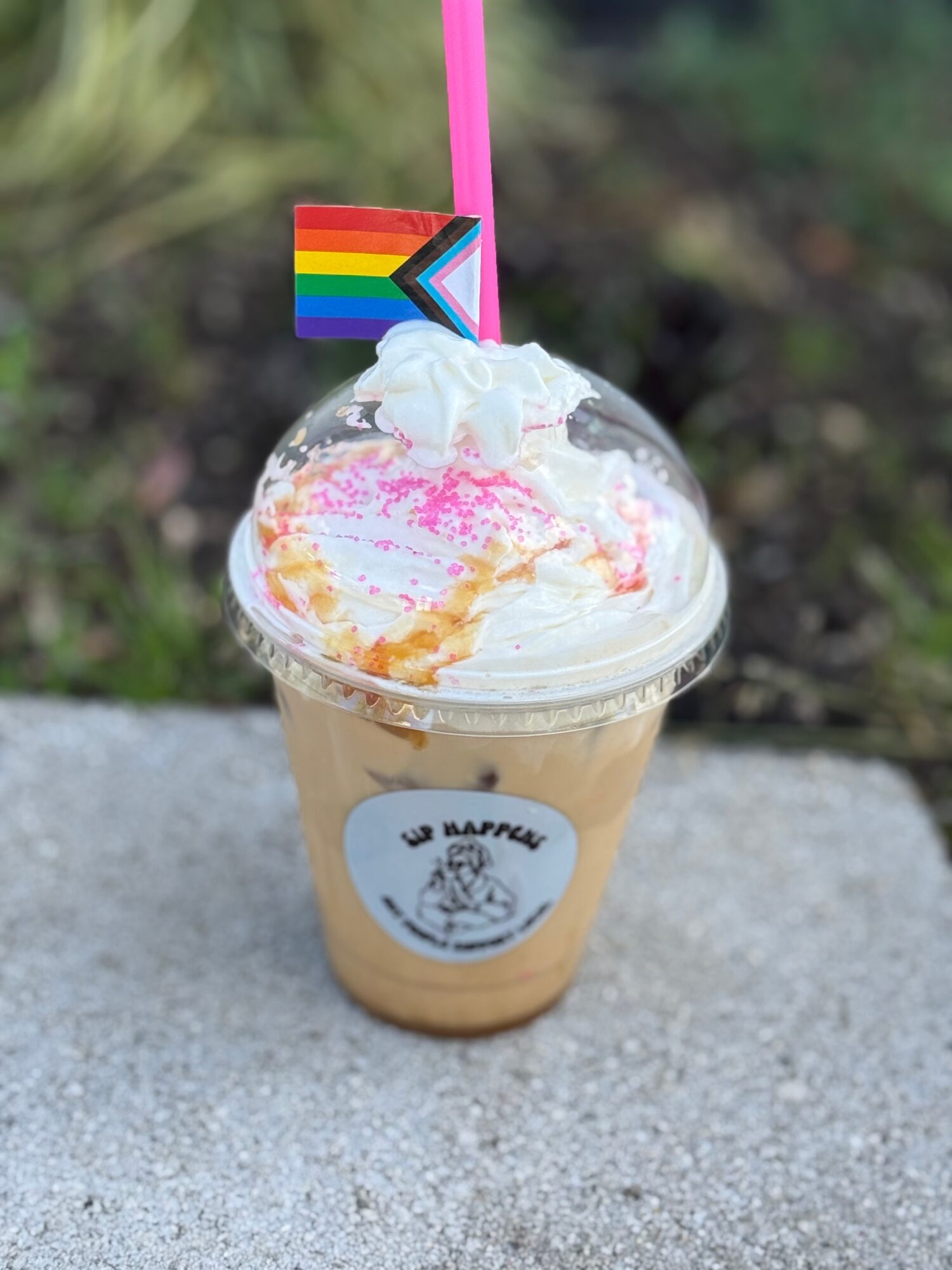 A cold beverage in a clear plastic cup with whipped cream, rainbow pride flag, and pink straw, outdoors on a gray surface.