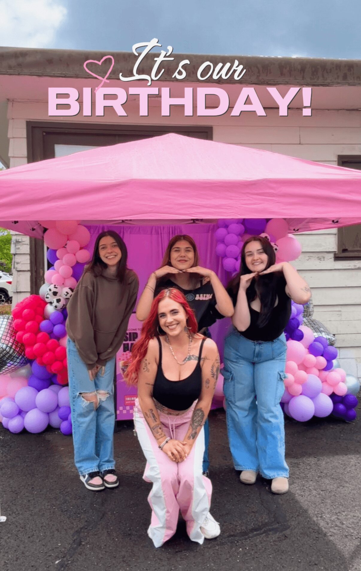 Four women stand in front of a pink birthday decoration with balloons and a pink canopy, smiling outdoors.