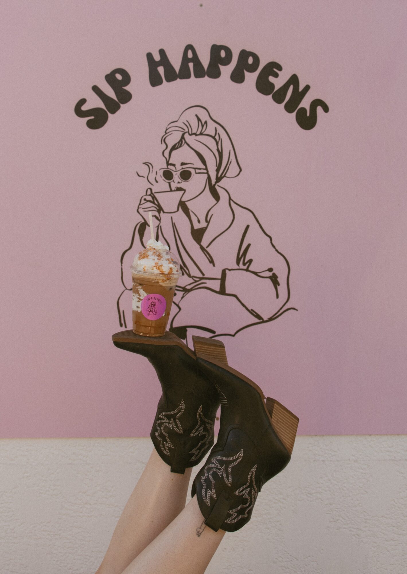 Line drawing of a woman with sunglasses, holding a drink, with text 'SIP HAPPENS' above, on a pink wall.