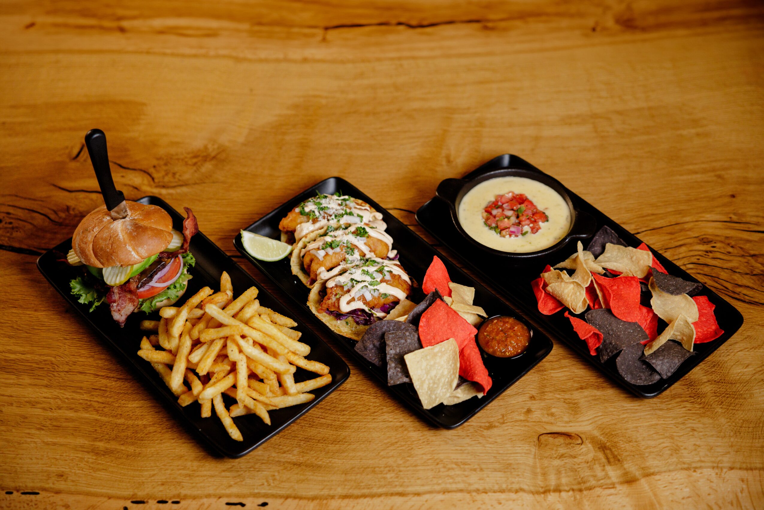 Three black trays with food on a wooden table: a burger with fries, a wrap with dipping sauce, and a bowl of soup with bread pieces.