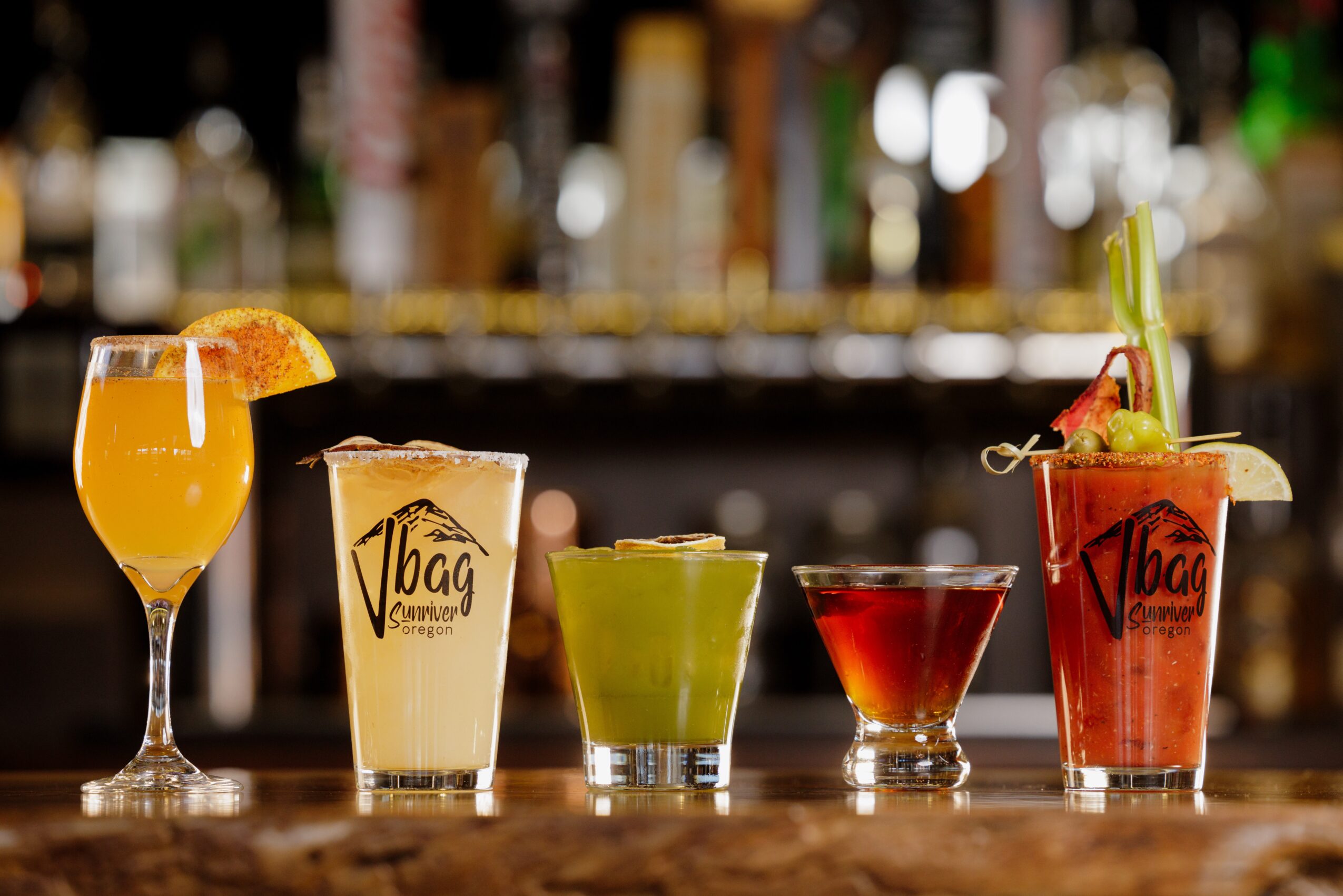 Five colorful cocktails on a bar counter with blurred bottles in background.