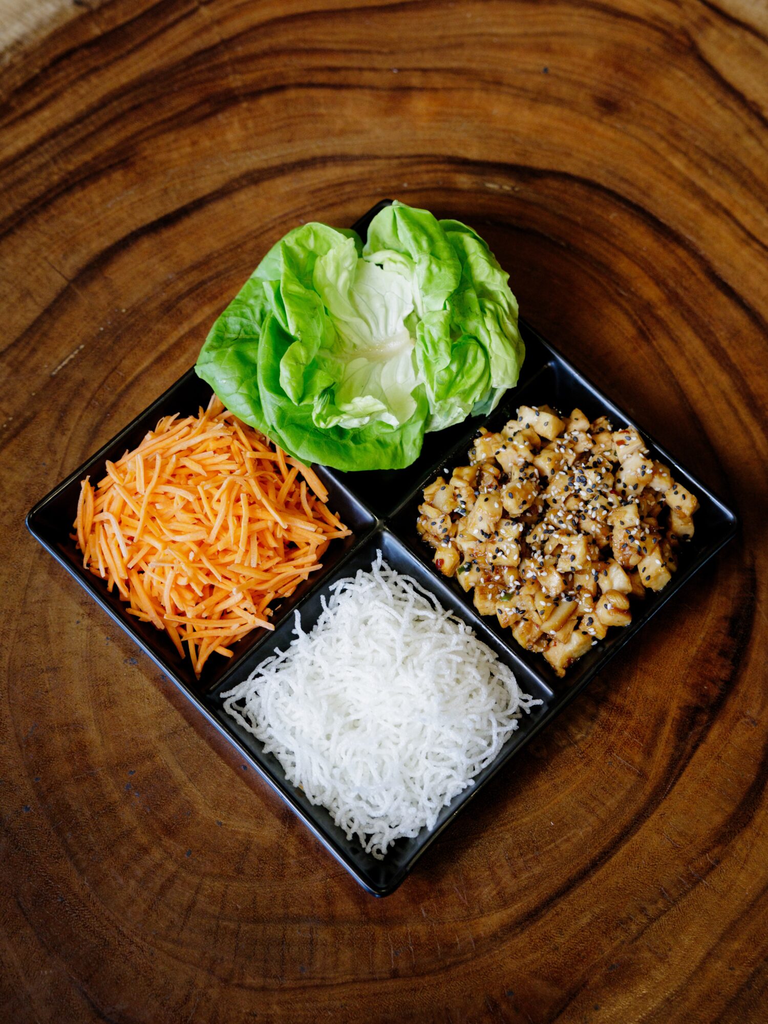 Four compartments with lettuce, shredded carrots, shredded white radish, and cooked ground meat on a black tray, on a wooden surface.