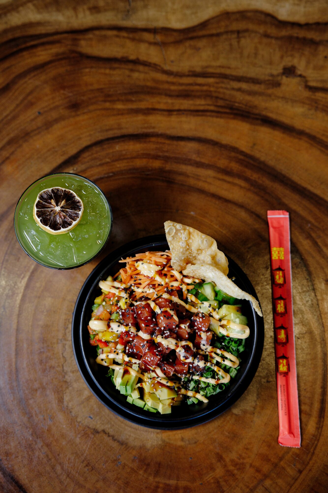 Bowl of salad with vegetables and dressing, a lime with a slice cut out, and a red chopstick on a wooden table.