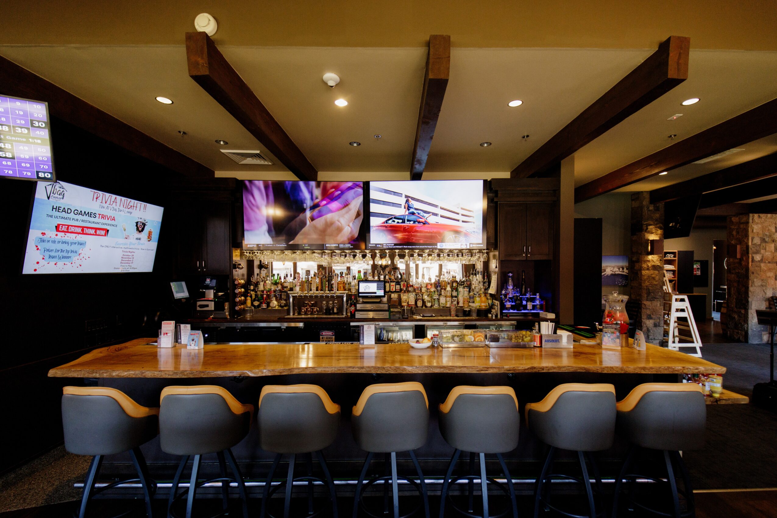 Bar with six chairs, three TV screens, bottles on shelves, and a dark interior with wooden beams.