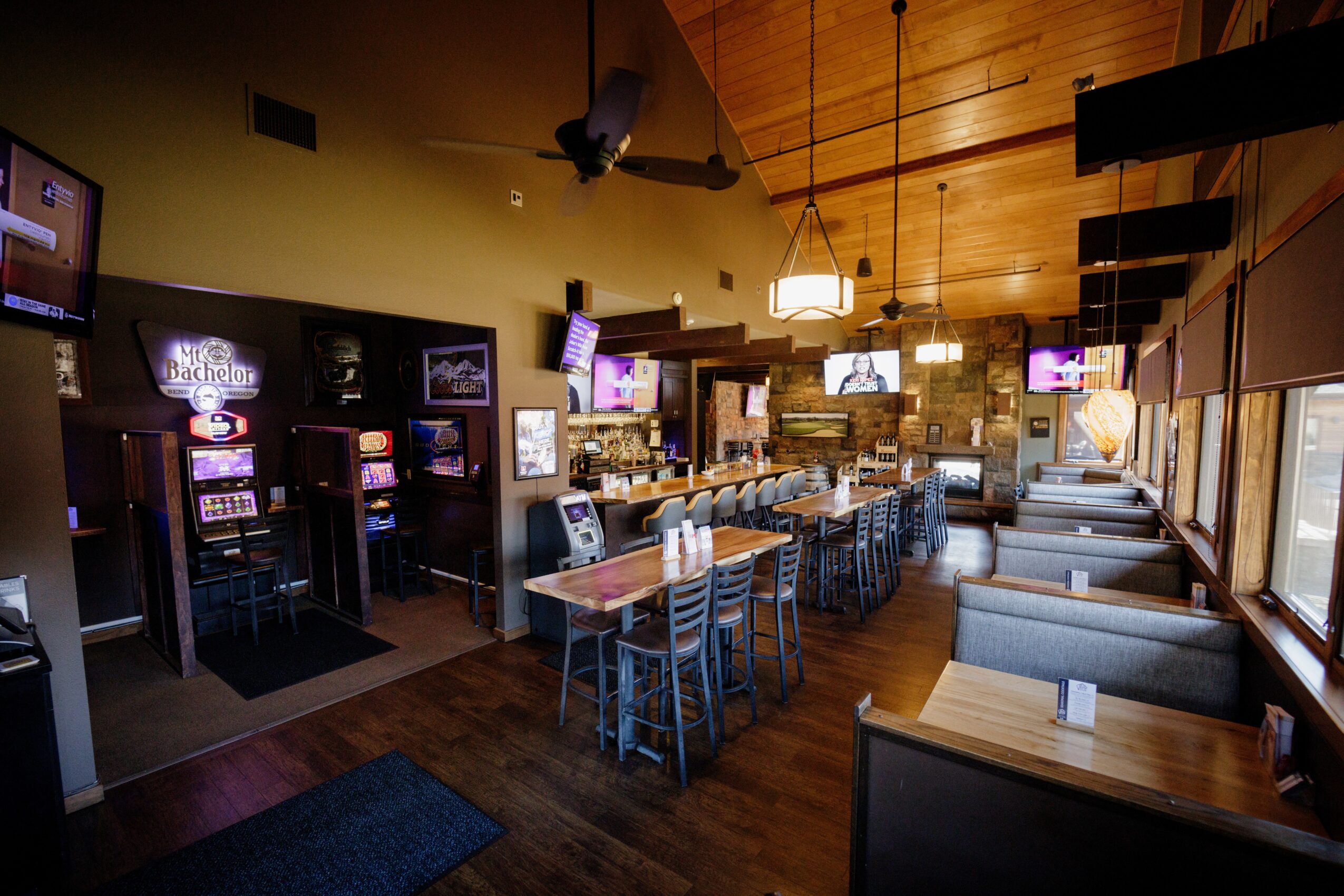 Empty restaurant with bar counter, tables, chairs, and booths, illuminated by hanging lights, with large windows on the right side.